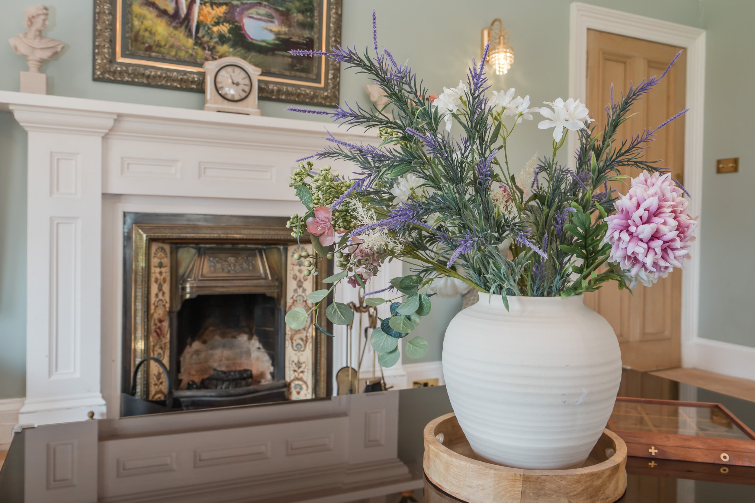 A white ceramic vase with a large bouquet of mixed flowers and greenery, sitting on a wooden stand in a living room with a fireplace, artwork, a clock, and decorative objects.
