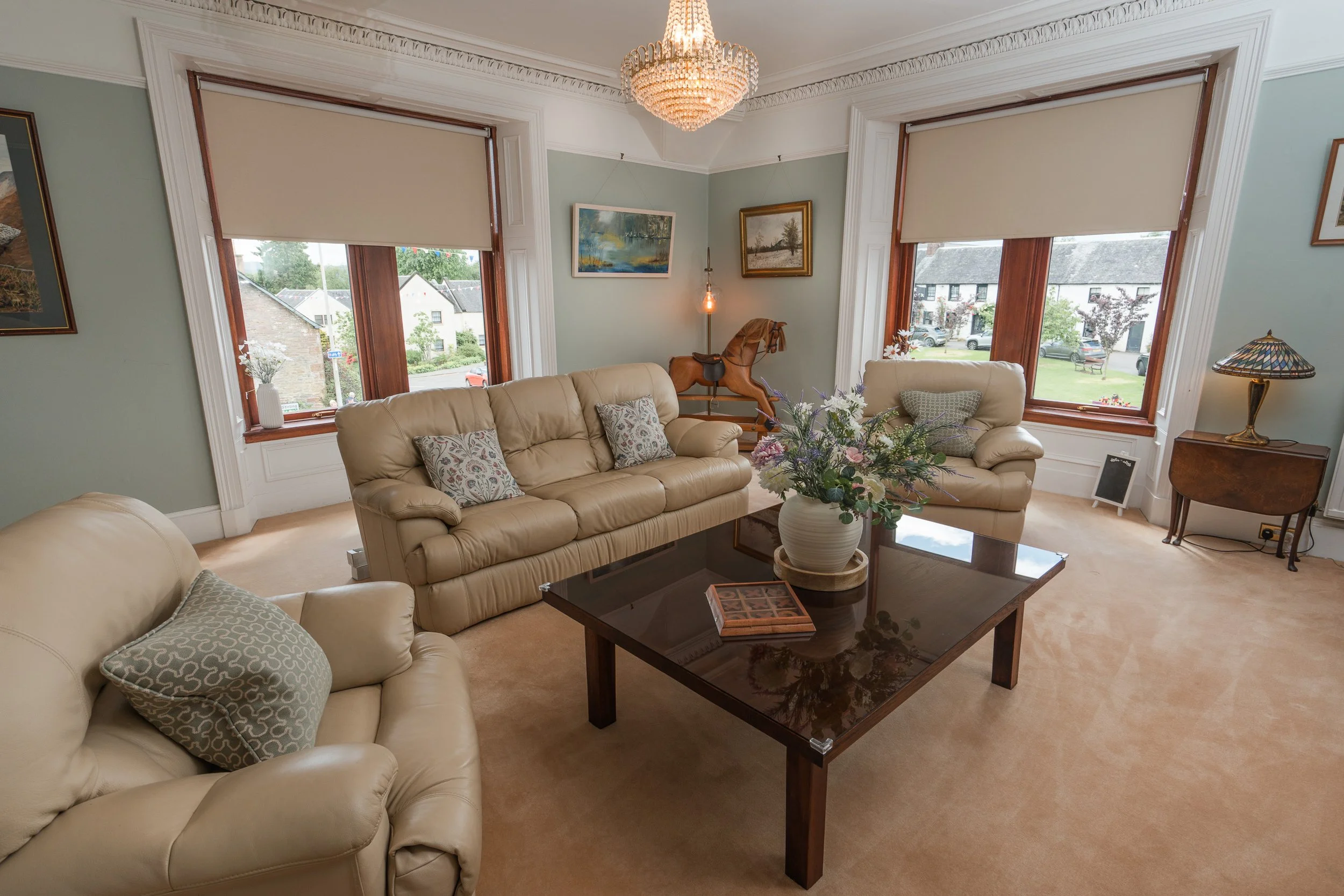 A living room with large windows, beige leather sofas with patterned pillows, a wooden coffee table with a vase of flowers, artwork on the walls, a vintage wooden horse sculpture, a Tiffany-style table lamp, and a chandelier hanging from the ceiling.