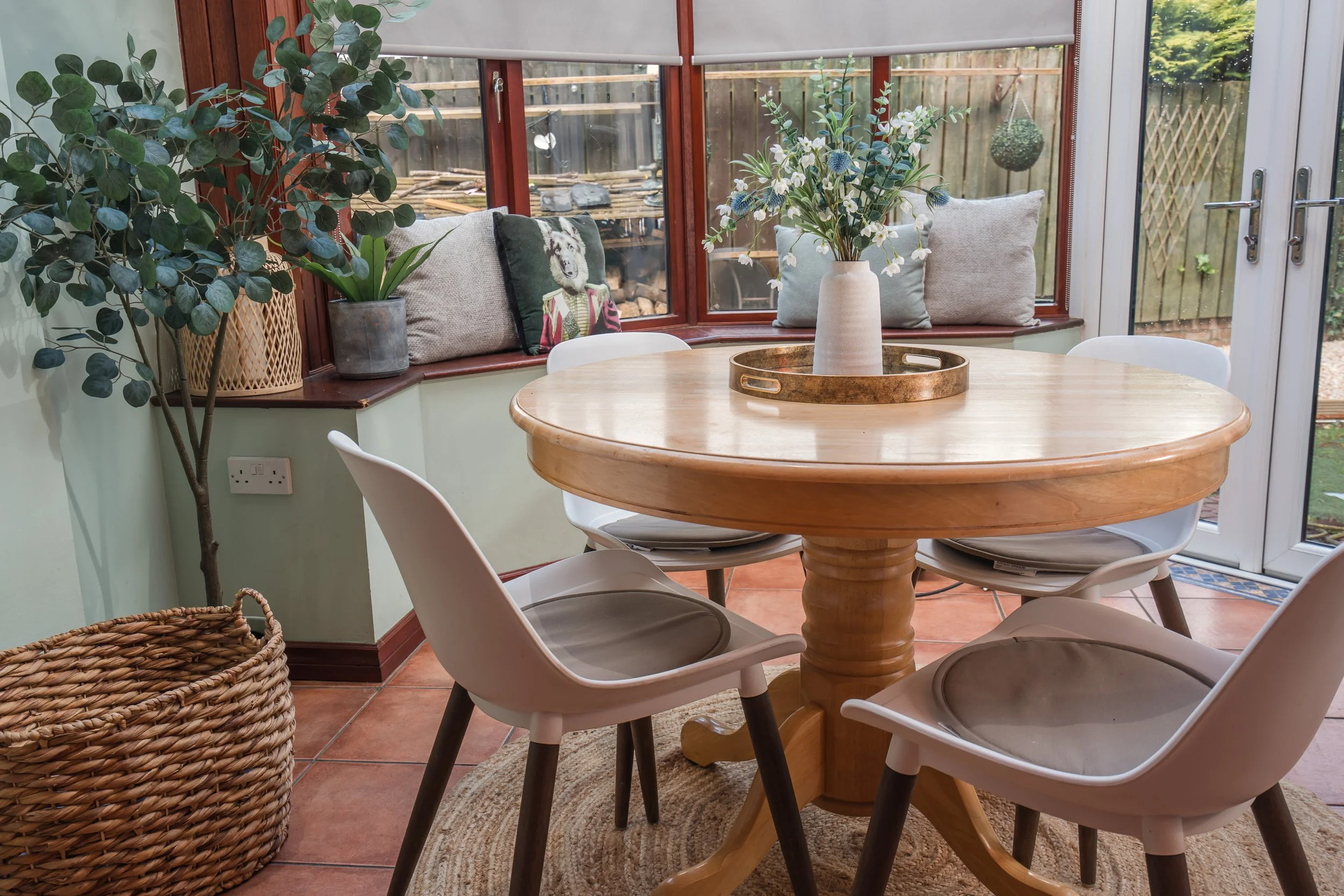 A cozy dining area with a round wooden table, white chairs, and a centerpiece vase of flowers. The space has large windows with cushions on the window ledge, and there are plants nearby, including a tall potted plant and a woven basket. Outside, a wo