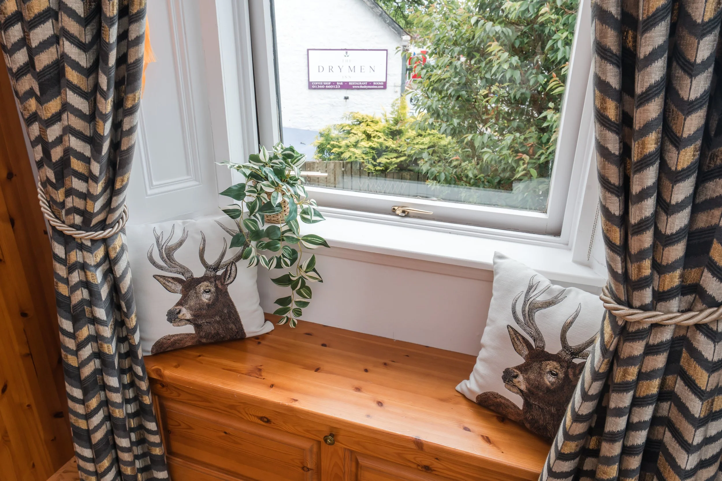 A cozy window seat with patterned curtains, a potted plant, and decorative pillows featuring a deer and a lion, outside view of greenery and a sign for 'The Drymen'