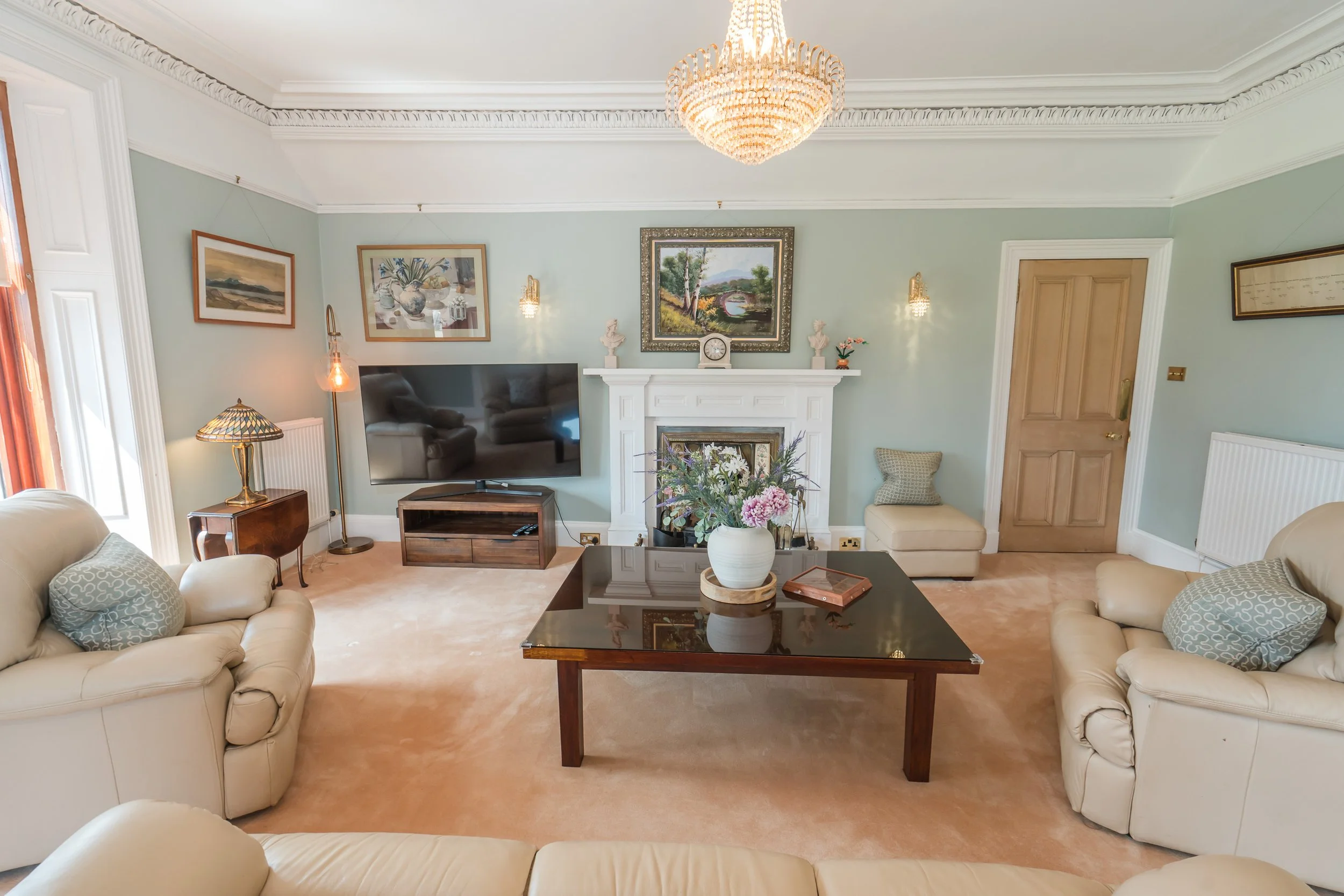 Living room with cream-colored sofas, a wooden coffee table with a vase of flowers, a flat-screen TV on a wooden stand, framed paintings on light mint green walls, a white fireplace mantel with decorative items, a chandelier, and a door on the right 