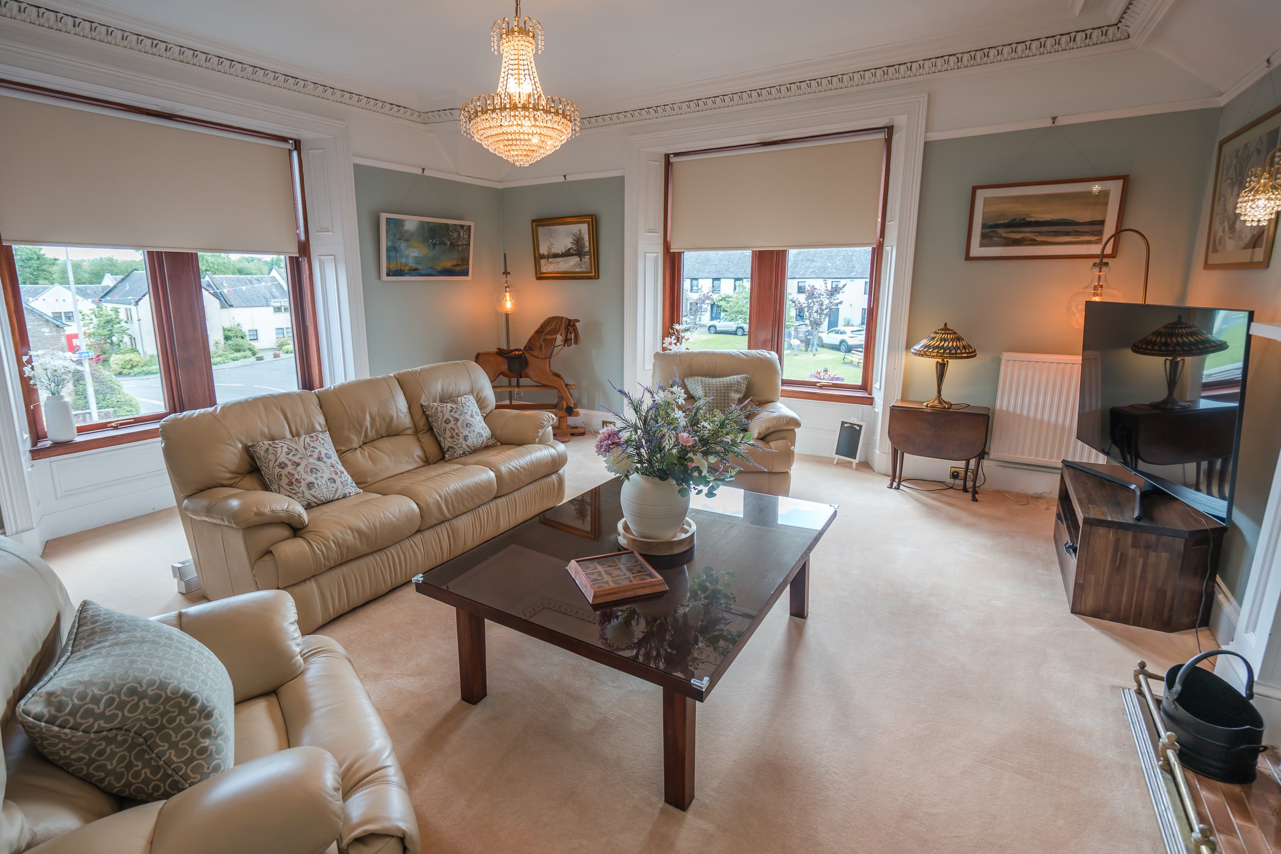 Living room with beige leather sofas, a wooden coffee table with a flower vase, large windows with blinds, paintings on walls, a chandelier, and a flat-screen TV.