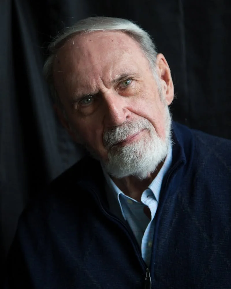 A portrait of an elderly man with a white beard and mustache, light-colored hair, and light eyes, wearing a dark jacket and a collared shirt, looking into the camera with a serious expression.