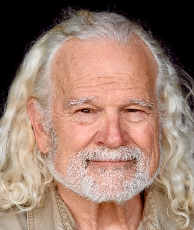 Close-up of an elderly man with long white curly hair, a full beard, and a gentle smile, wearing a beige shirt against a dark background.