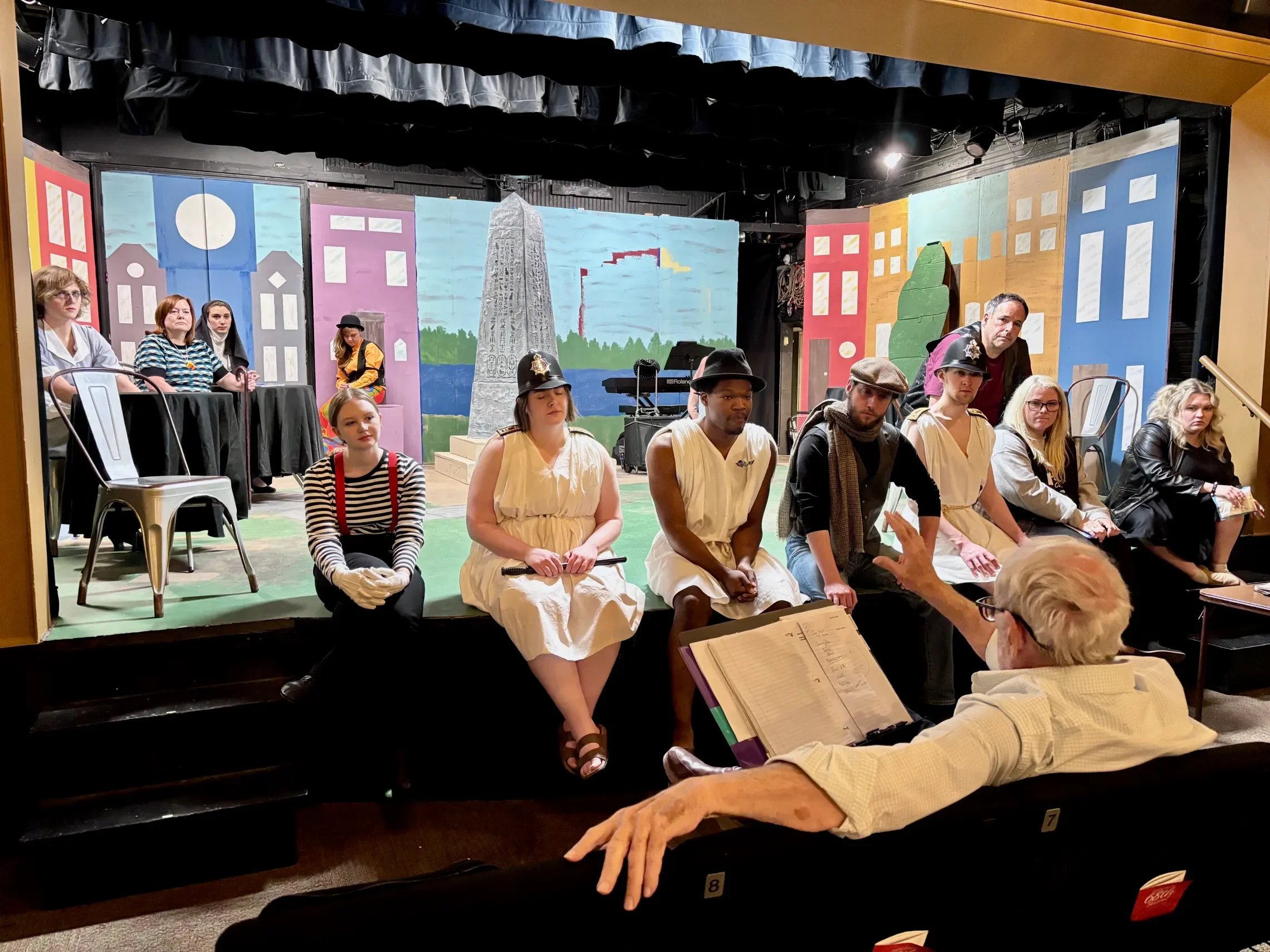People sitting on a stage with colorful cityscape backdrop including buildings and a monument, while a director gives notes.