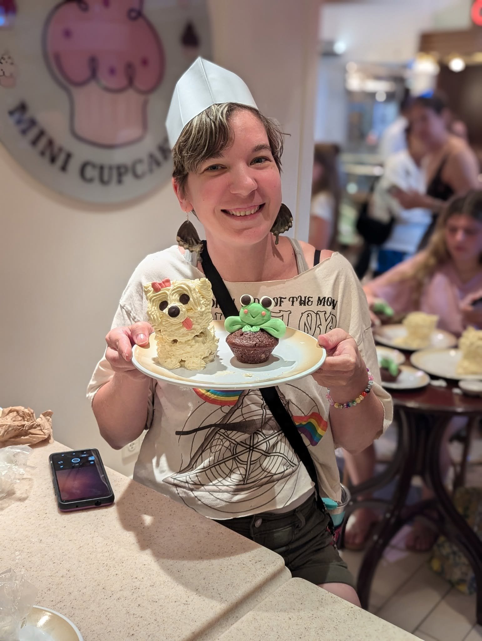 A woman with short hair, earrings, and a white paper hat holds a plate with decorated cupcakes that have animal faces, one resembling a dog and the other a frog. She is smiling in a bakery or café setting with other people in the background.