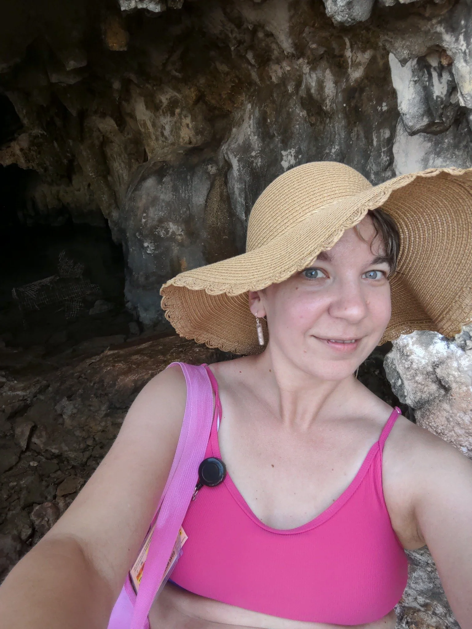 A woman with blue eyes and freckles taking a selfie outdoors, wearing a large straw sunhat and a pink tank top, with rocky terrain in the background.