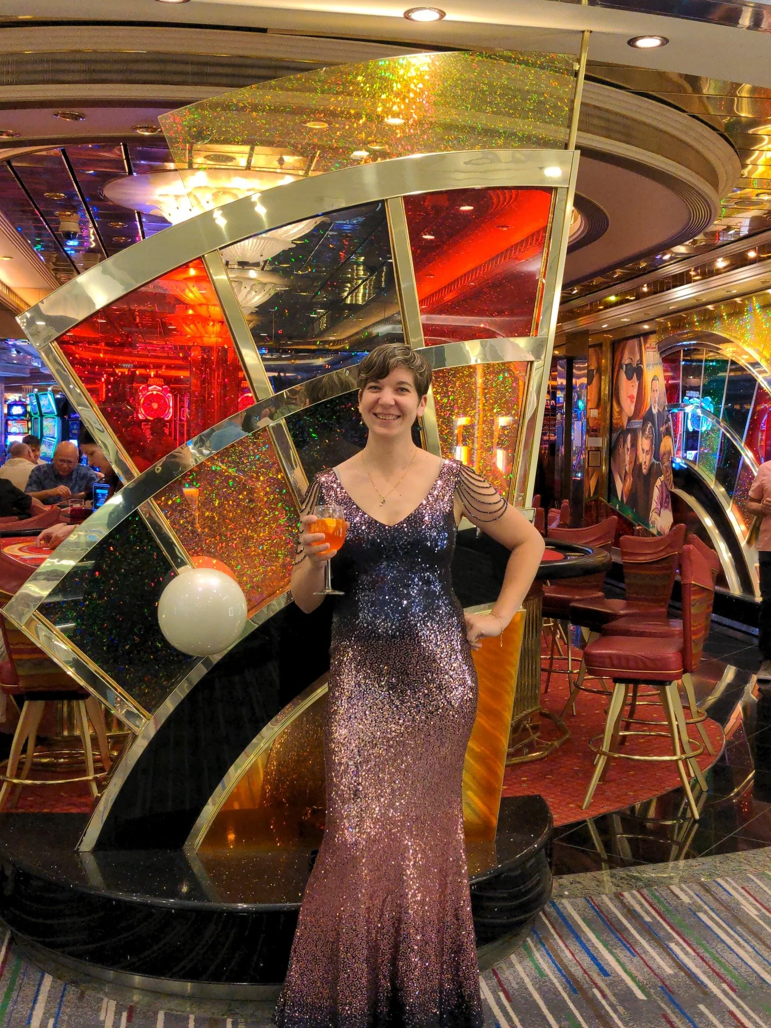 A woman in a sparkly, sequined, purple dress holding a drink inside a casino. She is smiling and standing near a shiny, decorative structure with colorful reflective panels. Thinking of a lively, glamorous setting with slot machines and casino decor in the background.