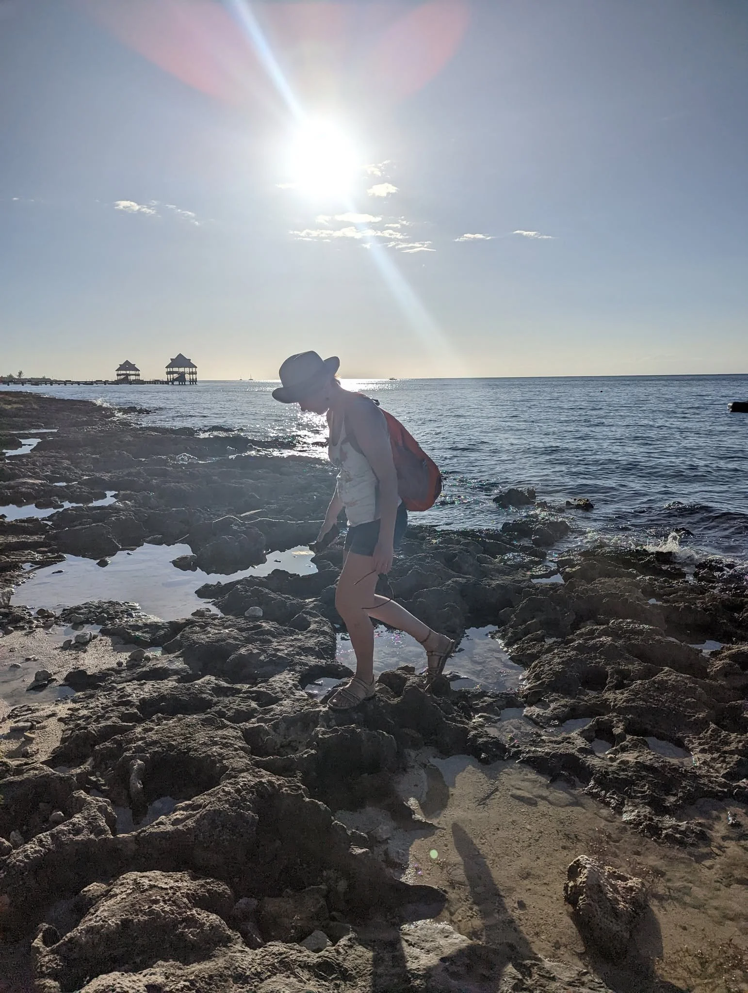 Person walking on rocks near the ocean with a clear sky, bright sun, and distant structures over water, wearing a hat and carrying a backpack.