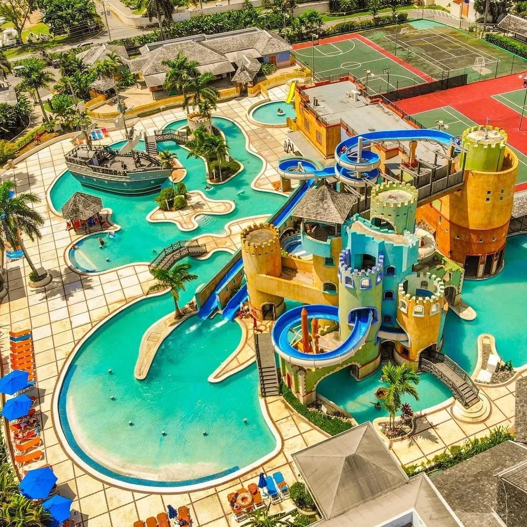 A colorful water park featuring a large splash pad with a ship-shaped water feature, a castle with blue and yellow turrets, and multiple water slides. Surrounding the water attractions are pools, palm trees, and lounge chairs. In the background, there are tennis courts and a small building.