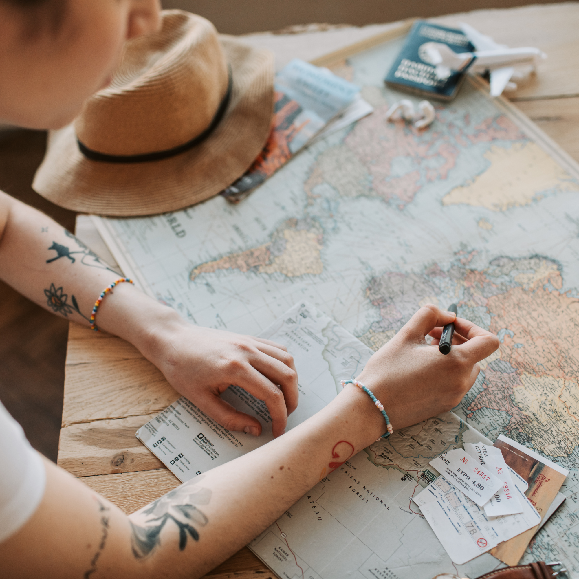 Person pointing at a map with airplane tickets and a notebook nearby, wearing a straw hat and colorful bracelets.