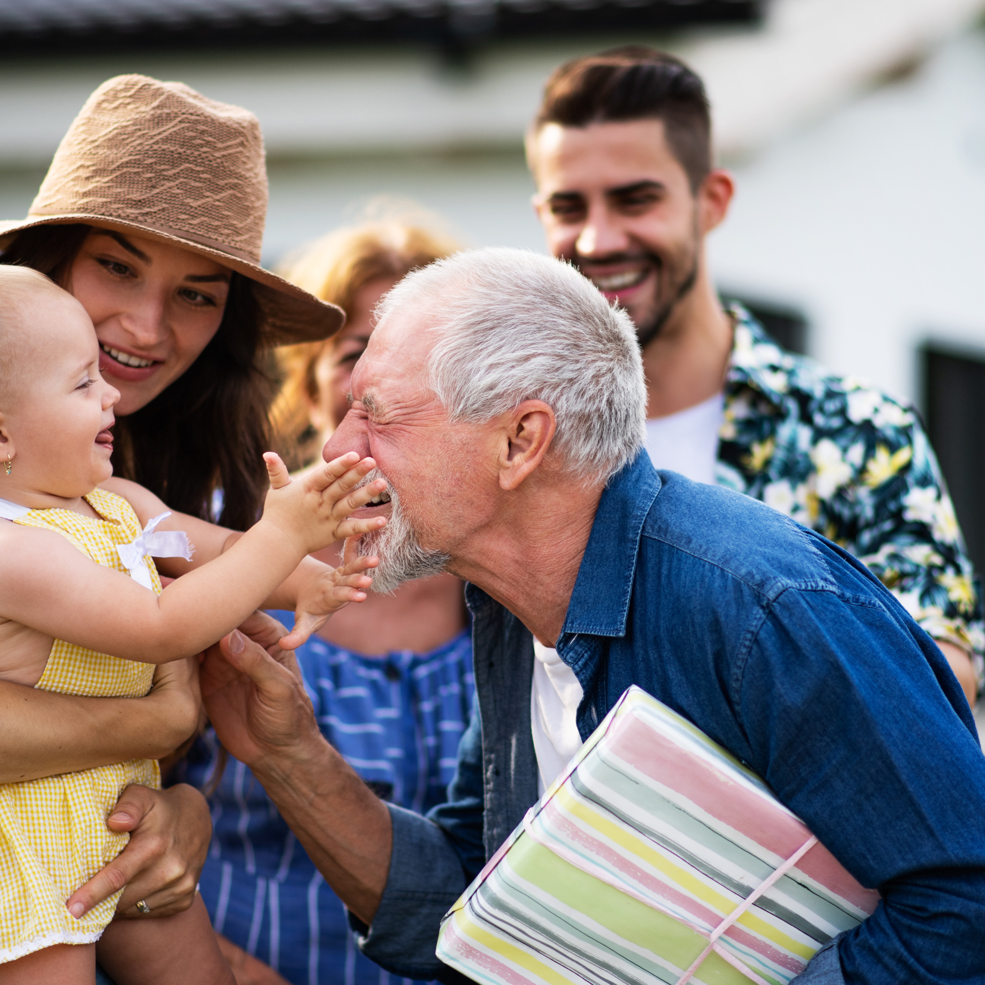A group of people, including a woman wearing a tan hat and a young girl in a yellow dress, smiling and interacting at a gathering during daytime. A man with dark hair and a floral shirt is in the background, and someone with white hair is in the foreground holding a wrapped gift with colorful stripes.