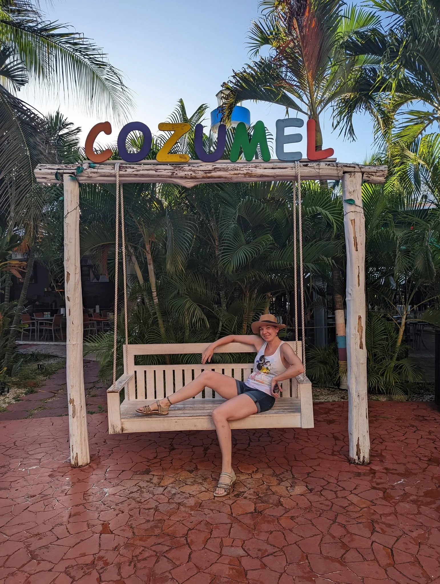 A woman resting on a wooden swing in a tropical setting with palm trees. There is a colorful sign on top that says 'COZUMEL' in multicolored letters. She is smiling, wearing a hat, shorts, and sandals, and appears relaxed and happy.