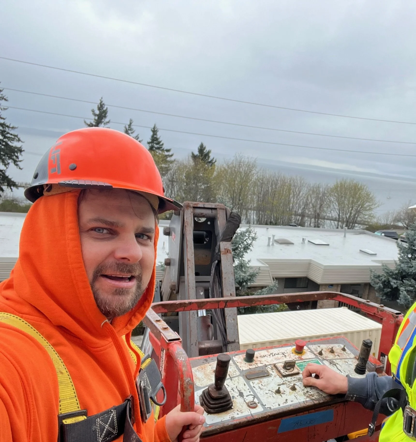 Mark wearing an orange safety jacket and hard hat operating machinery controls outdoors with cloudy sky and trees in the background.