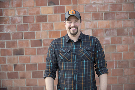Morgan in front of a brick wall, smiling, wearing a plaid shirt and a cap with 'FR' on it.