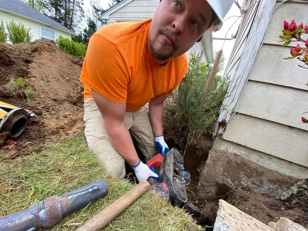 Morgan in a construction site digging near the foundation of a house. There are tools including a pipe cutter and a sledgehammer nearby, and a large hole is dug next to the house's exterior wall with visible soil.