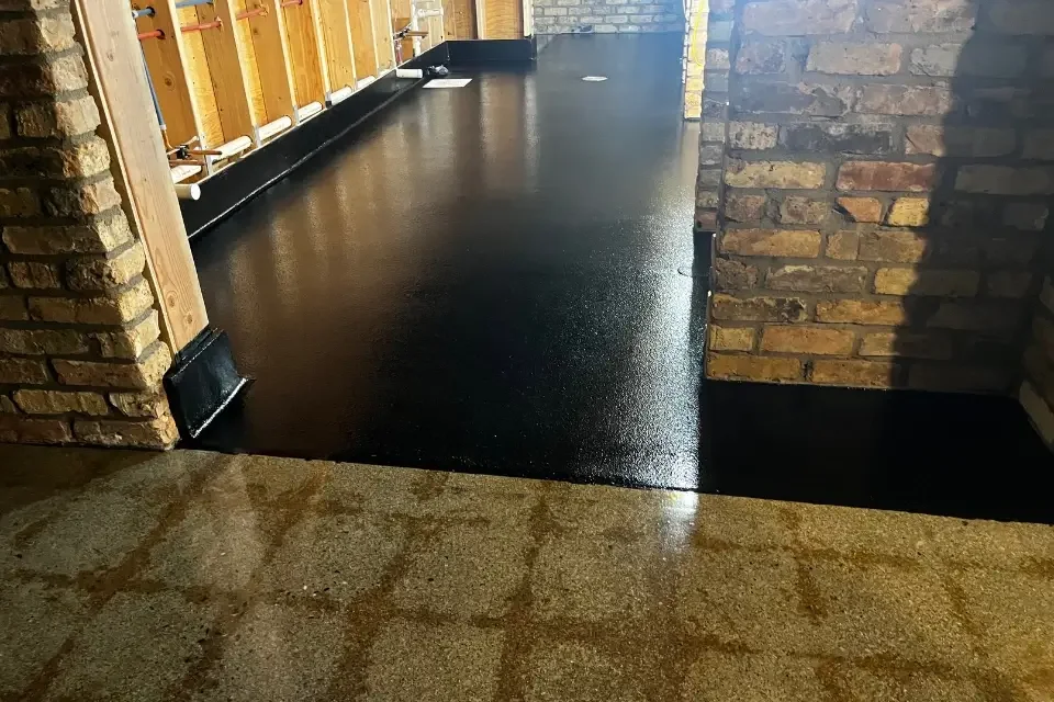 A seamless, glossy black waterproof floor coating applied in a basement renovation next to brick walls and exposed framing, highlighting durable moisture protection for damp environments.