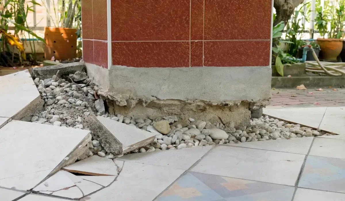 Damaged concrete foundation around a tiled structure with crumbling edges, broken floor tiles, and exposed gravel.