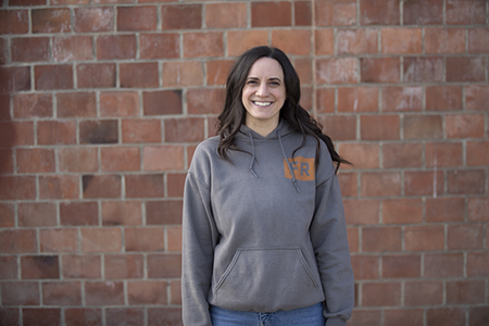 Kelsey wearing a gray hoodie and standing in front of a brick wall.