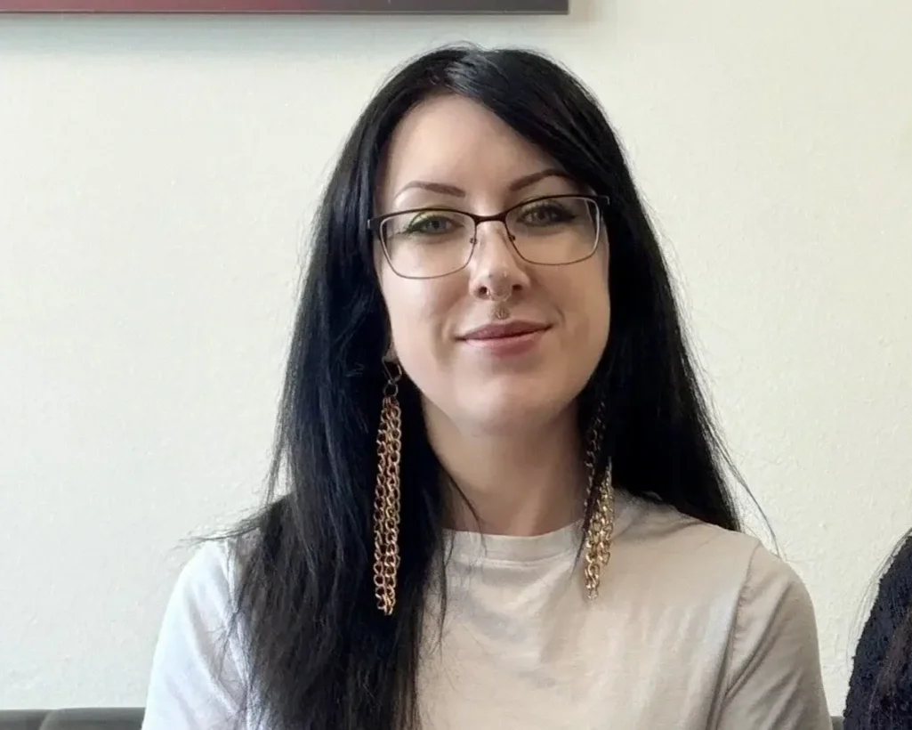 A woman with long black hair and glasses smiling at the camera, wearing a white shirt and long chain earrings, indoors with a plain wall in the background.