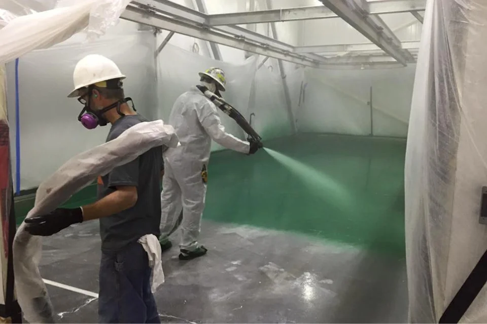 A technician wearing protective gear and a respirator sprays a green polyurea or epoxy coating onto a large indoor floor, demonstrating professional application techniques that maximize flooring lifespan.