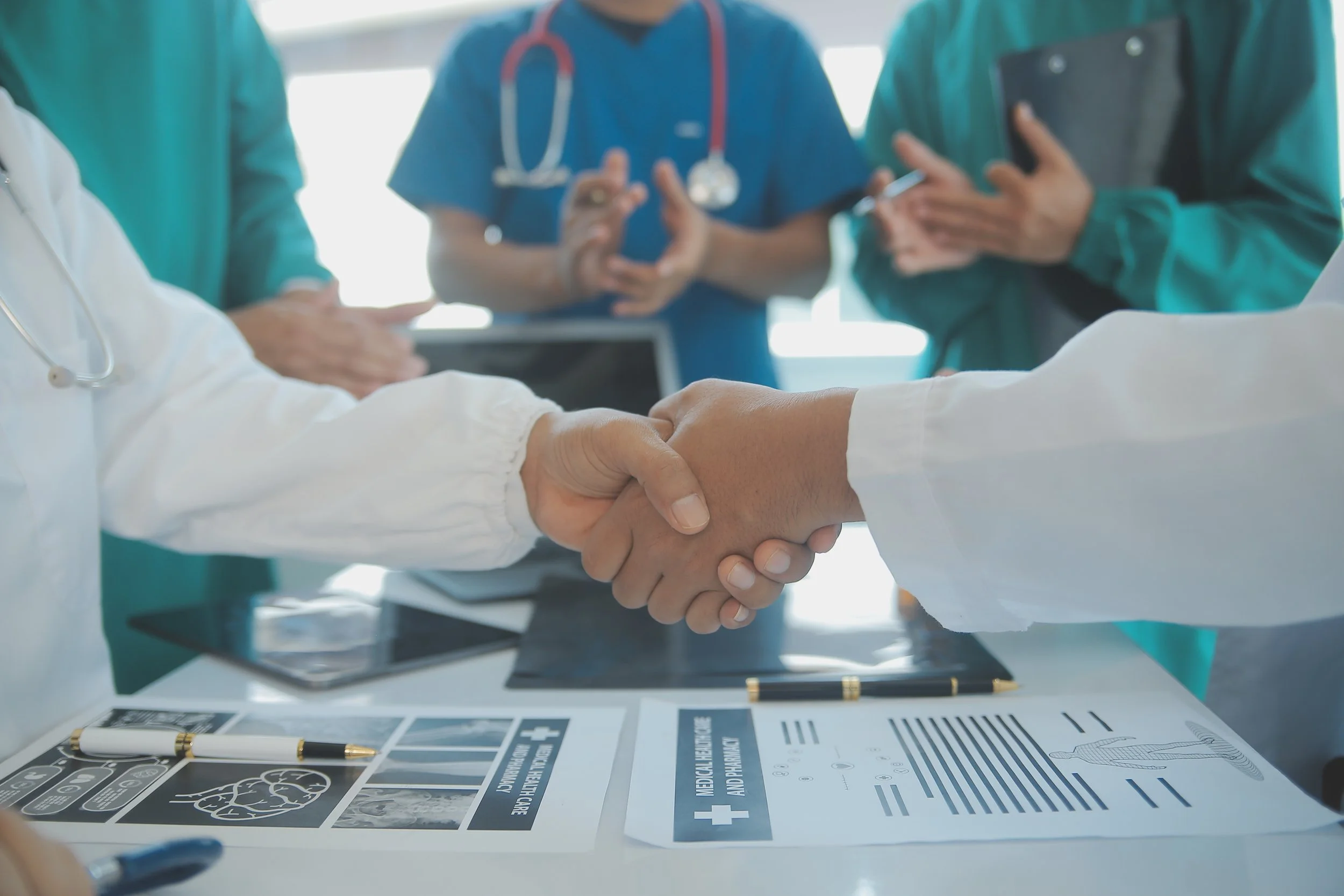 Close-up of a handshake between two medical professionals in scrubs, with a group of colleagues in the background, and a table with medical diagrams and pamphlets in the foreground.