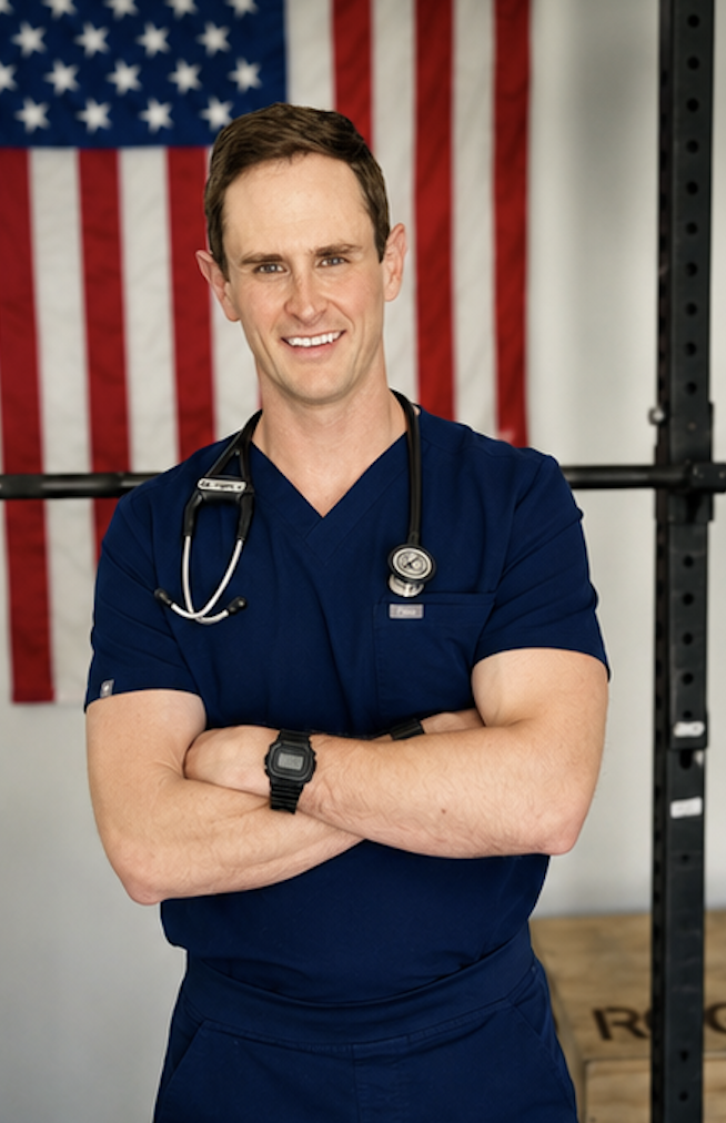 A male healthcare professional in blue scrubs with a stethoscope around his neck, standing with arms crossed in front of an American flag.
