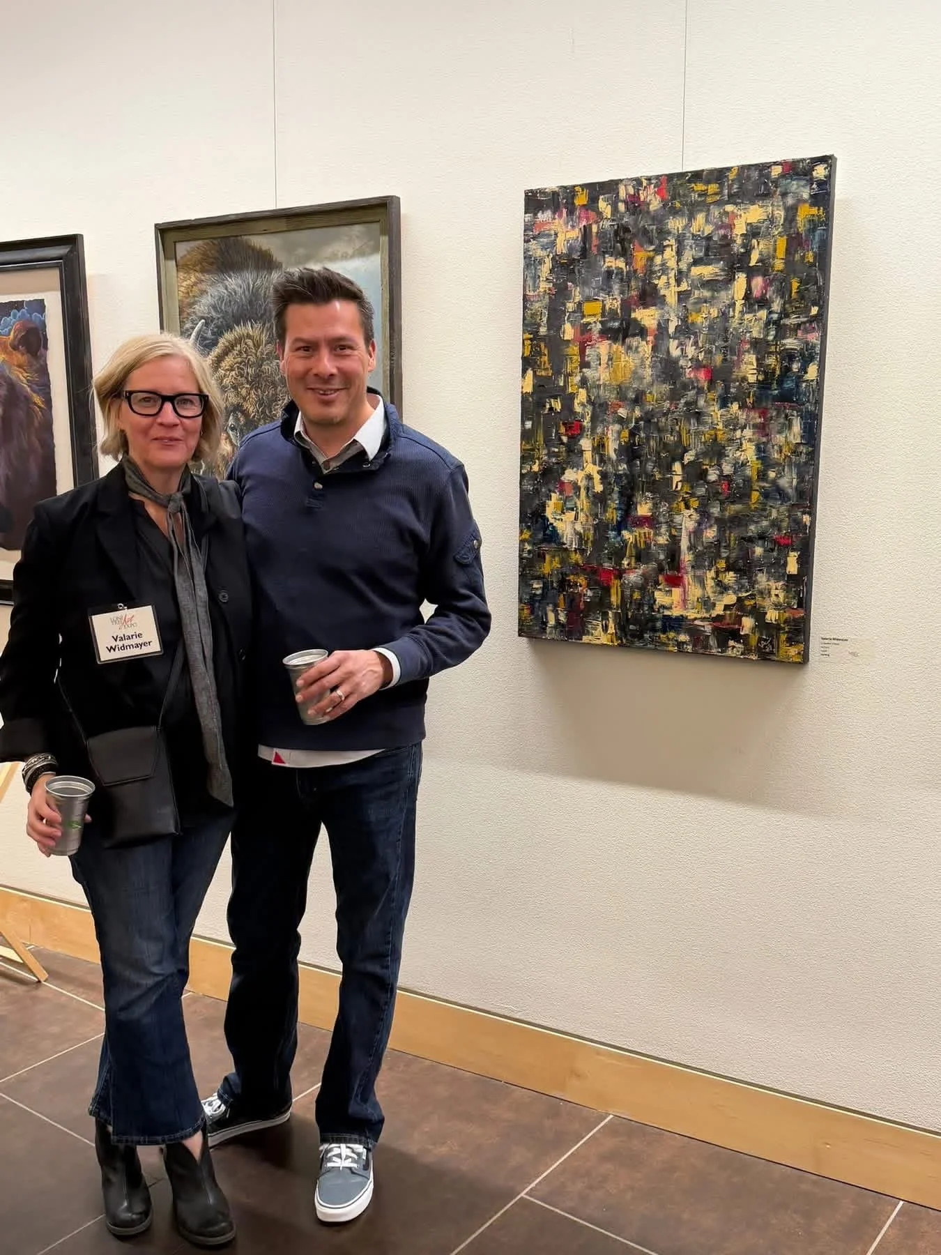 A woman and a man are standing together in an art gallery, smiling at the camera. The woman wears glasses, a black jacket, and has a name tag that reads Valerie Widmayer. The man is in a dark blue sweater and jeans, holding a drink. Behind them are various artworks, including a detailed animal portrait and an abstract painting.