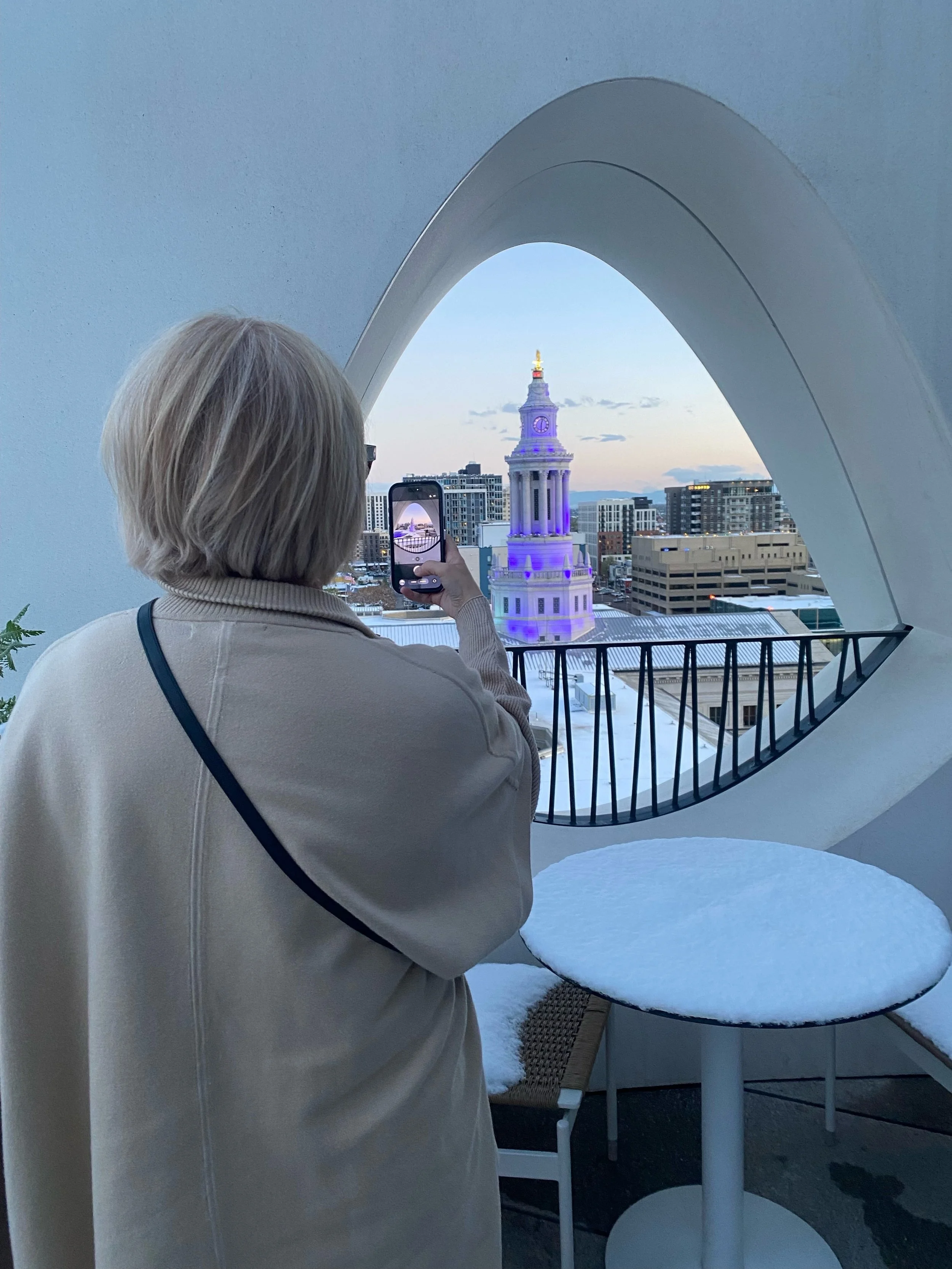 A woman with blonde hair taking a photo of a historic clock tower with a smartphone. The scene is viewed through a unique architectural opening with a smooth, curved frame. The cityscape shows multiple buildings, and the clock tower is lit with purple and yellow lights.