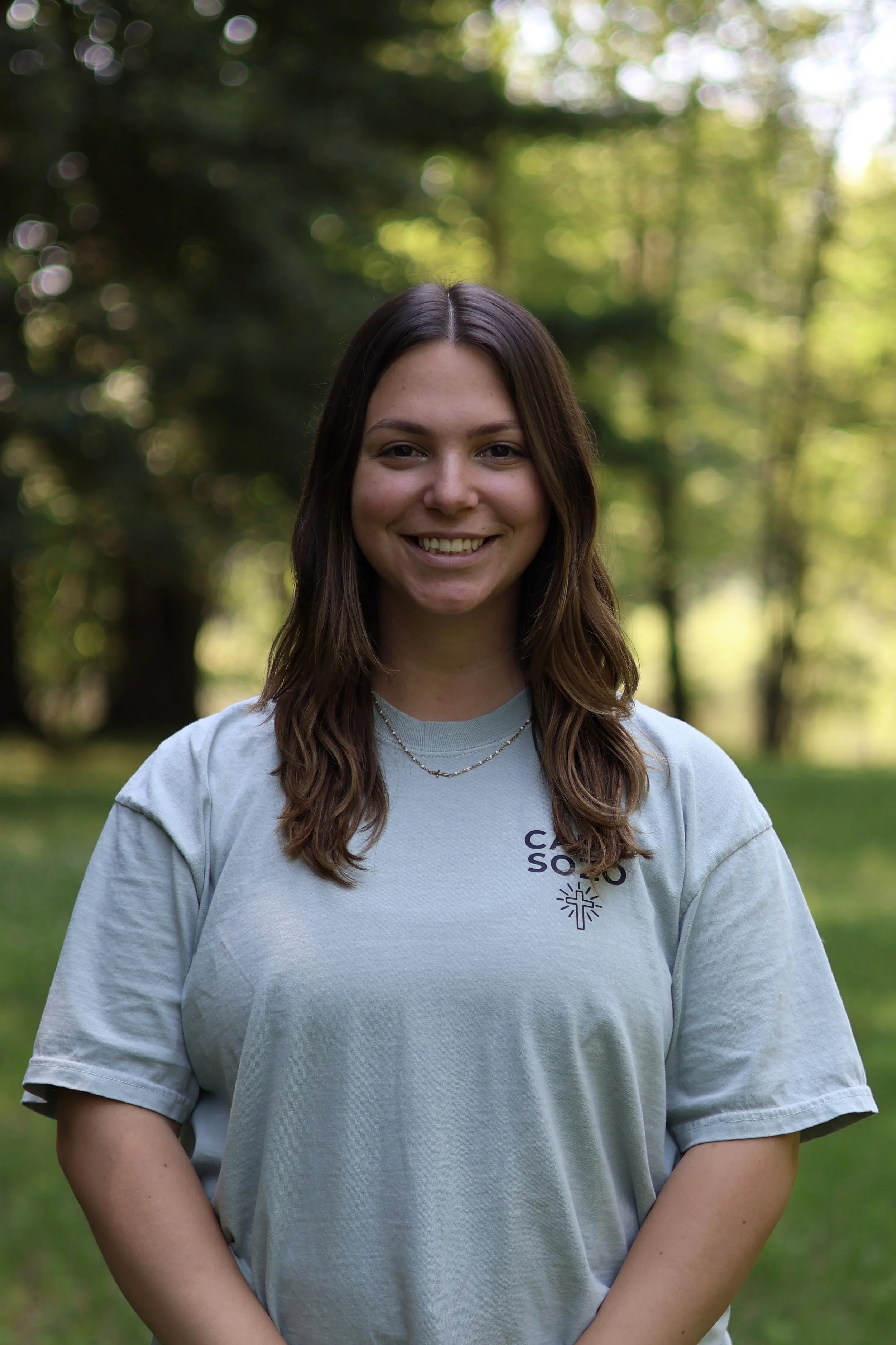 A young woman with long brown hair smiling outdoors on a sunny day, wearing a light grey T-shirt with text and a logo on it, in a natural environment with trees and green foliage in the background.