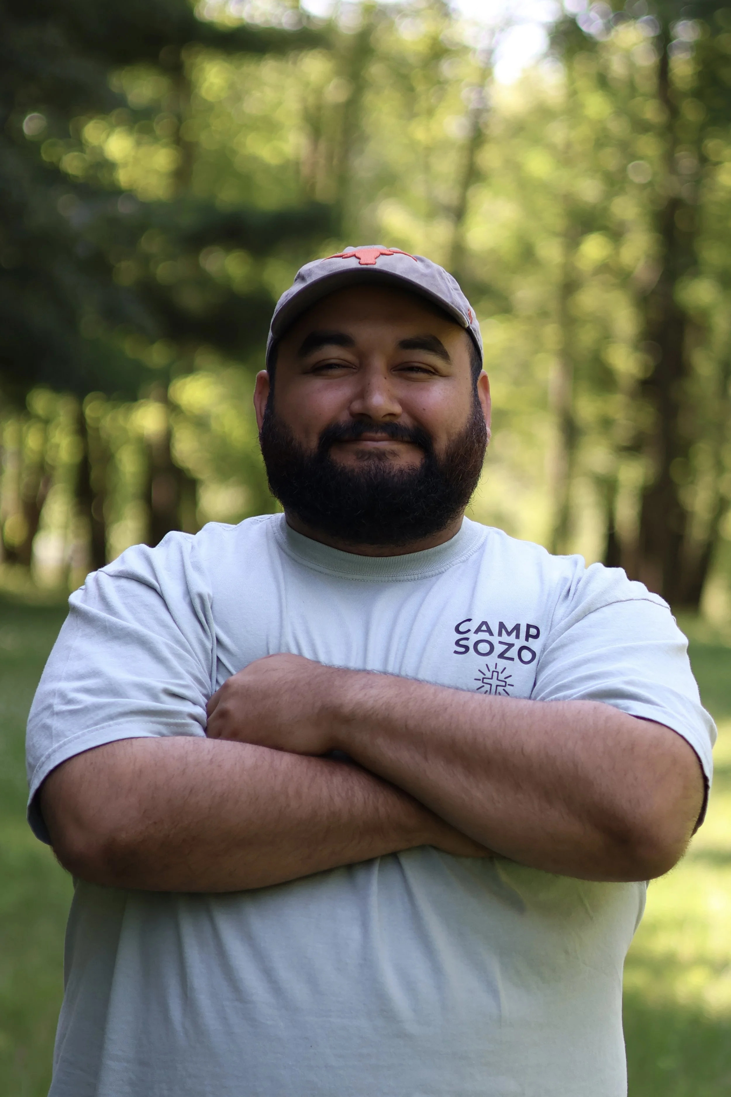 A man with a beard and mustache wearing a cap and a t-shirt with 'Camp Sozo' logo, standing outdoors in a park with green trees in the background, smiling with arms crossed.