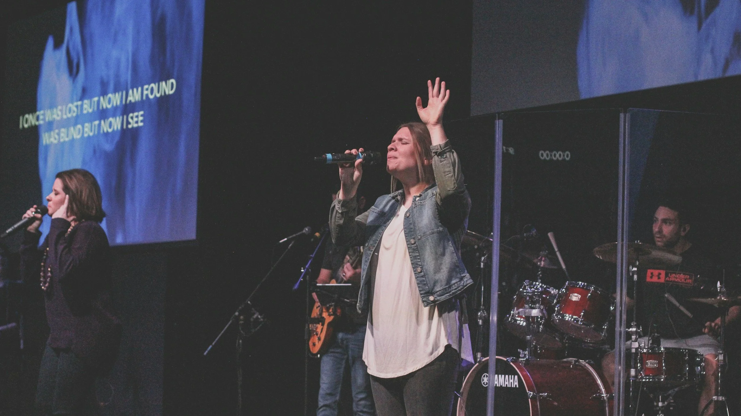 A woman with long hair sings into a microphone with her eyes closed, wearing a denim jacket and a white dress, during a live musical performance on stage. Other musicians, including a drummer and a keyboardist, are visible in the background.