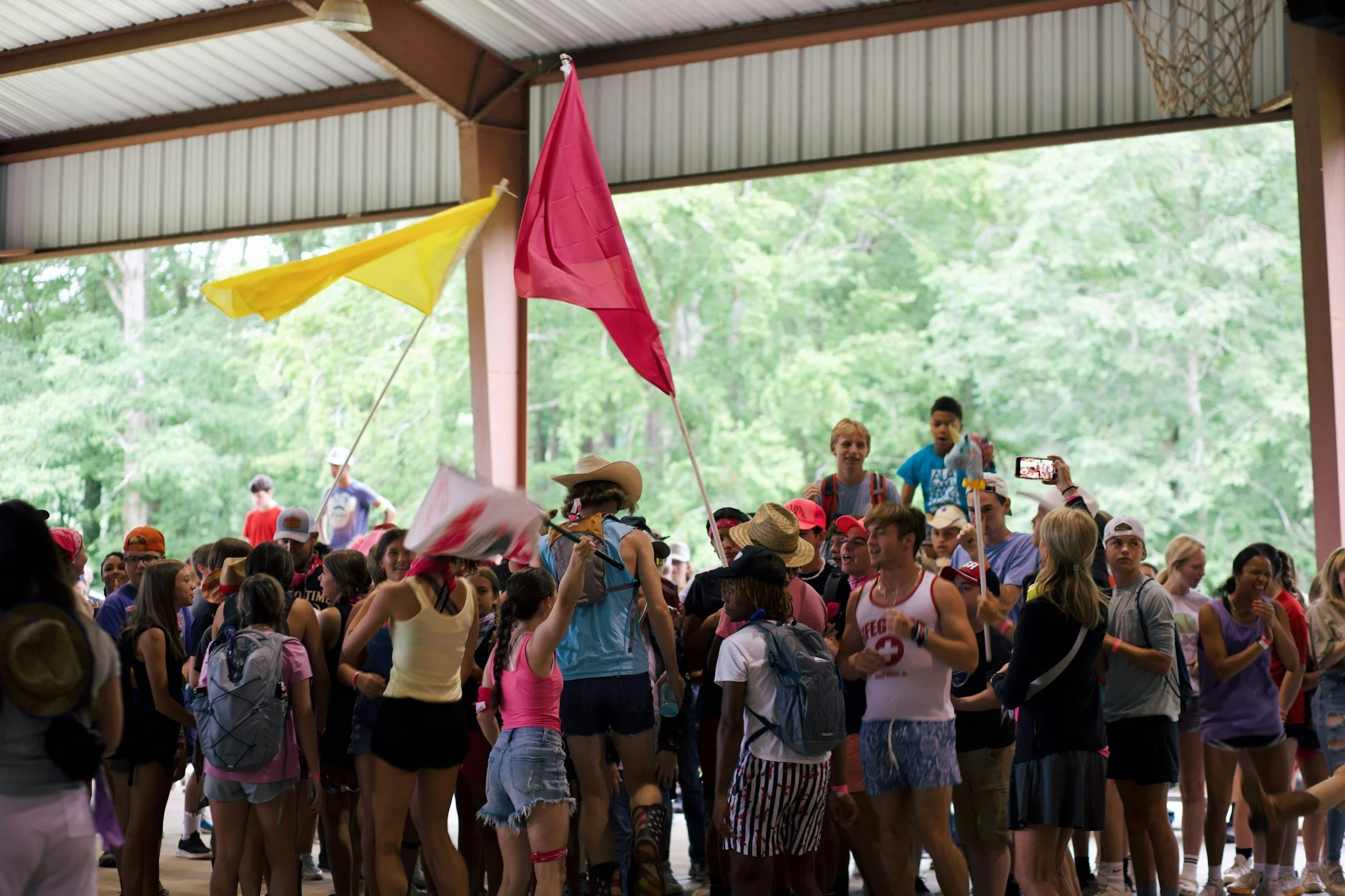 A large group of people gathered inside a covered outdoor pavilion, many wearing hats and casual clothing, with some holding flags and taking photos. The background features trees and greenery.