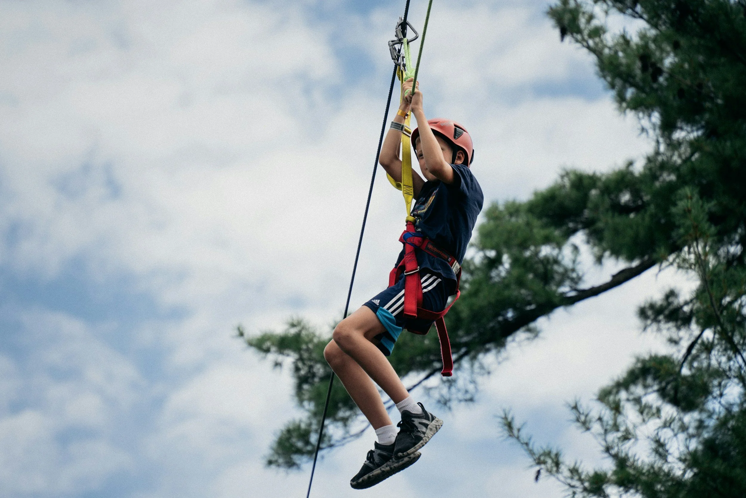 A boy wearing a red helmet and harness is ziplining through a forested area against a cloudy sky.