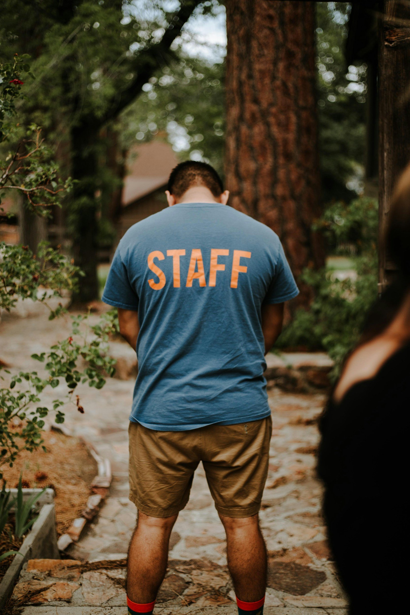 A person with short dark hair wearing a blue T-shirt with the word 'STAFF' in orange letters on the back, tan shorts, and red socks stands outdoors on a stone pathway with plants and trees around.
