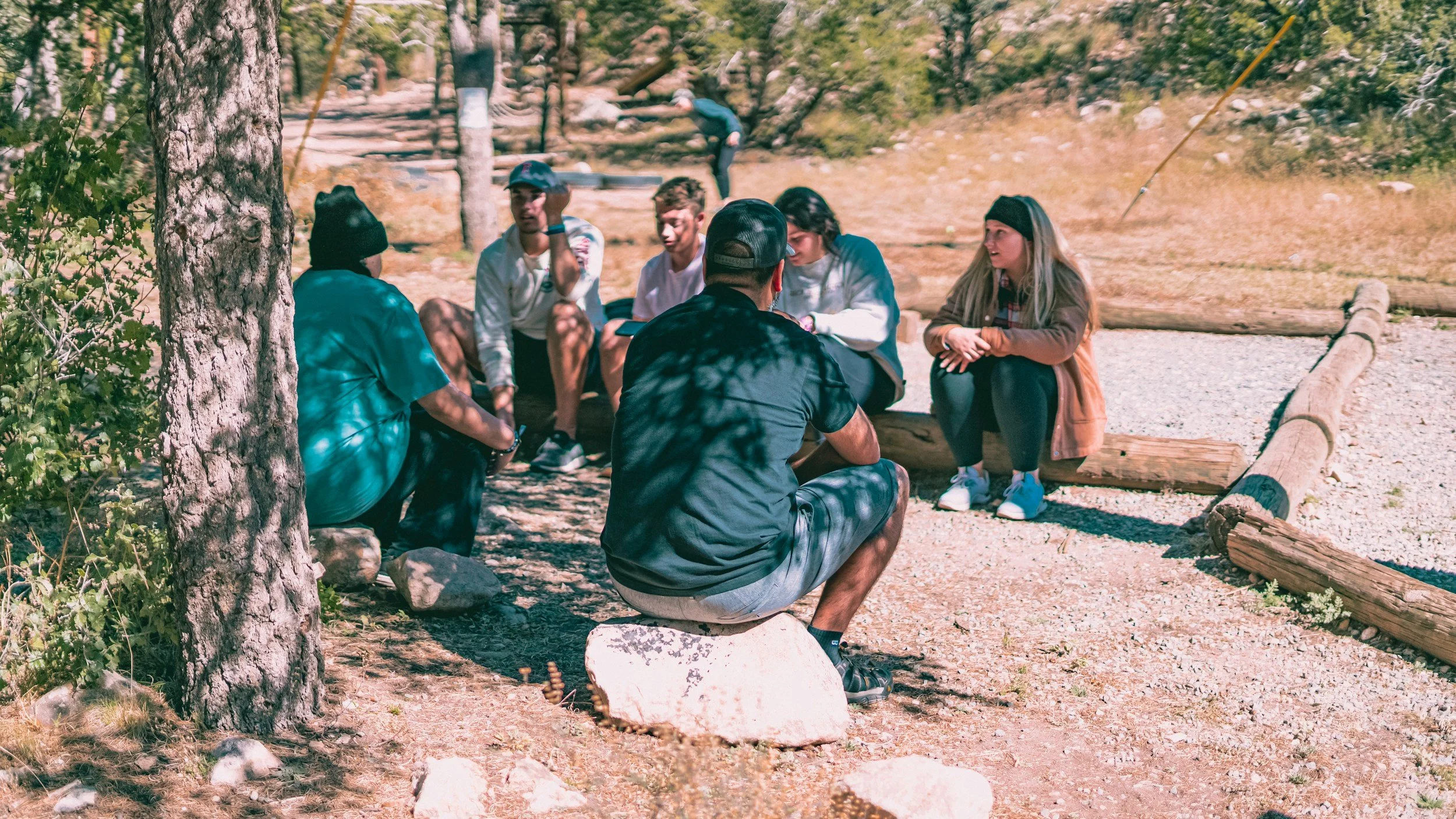A group of seven young people sitting outdoors in a wooded area, engaged in conversation and enjoying nature, with a couple of people in the background.
