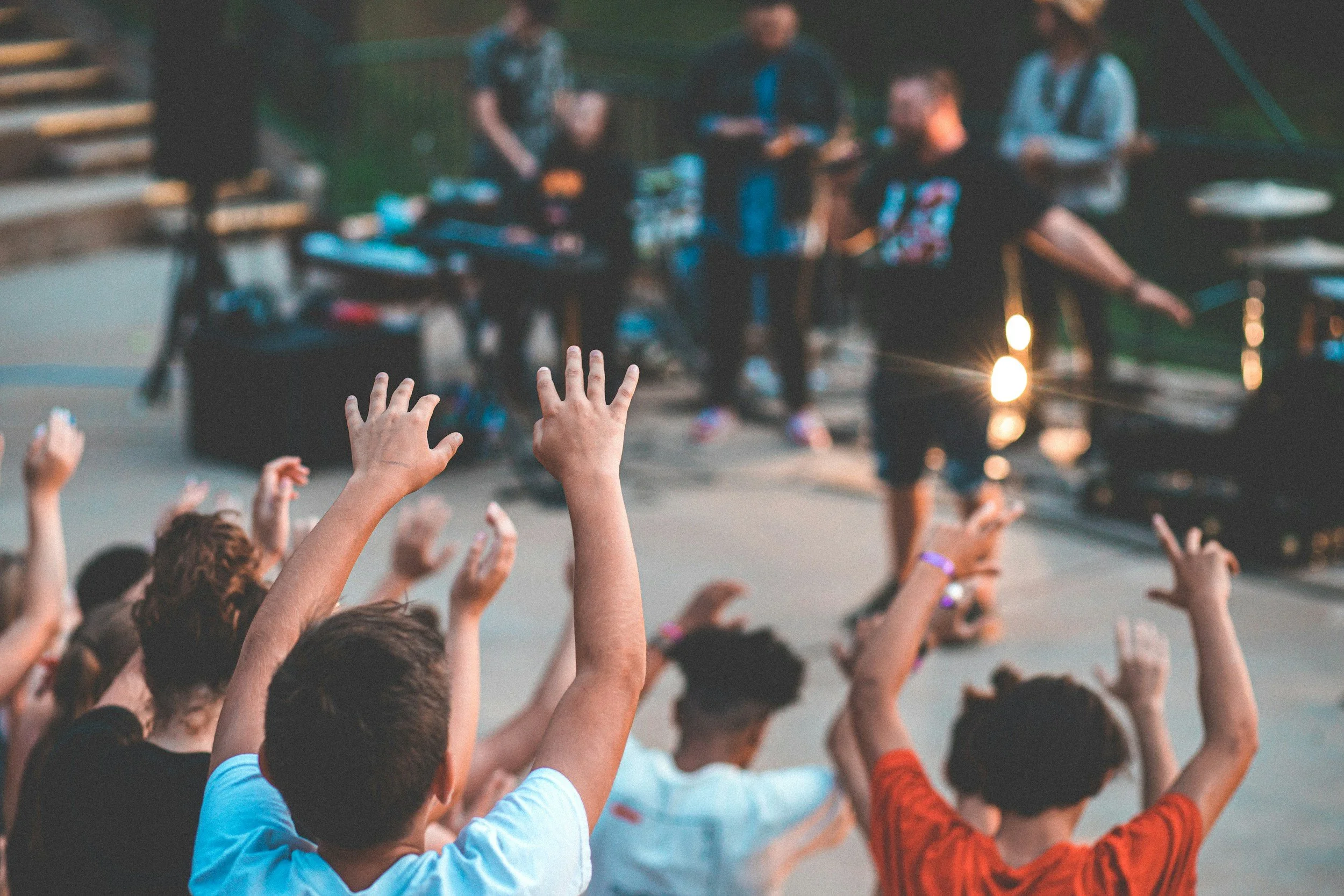 A crowd of people reaching their hands in the air as a music or entertainment event takes place outdoors, with a blurry group of performers or speakers on stage in the background.