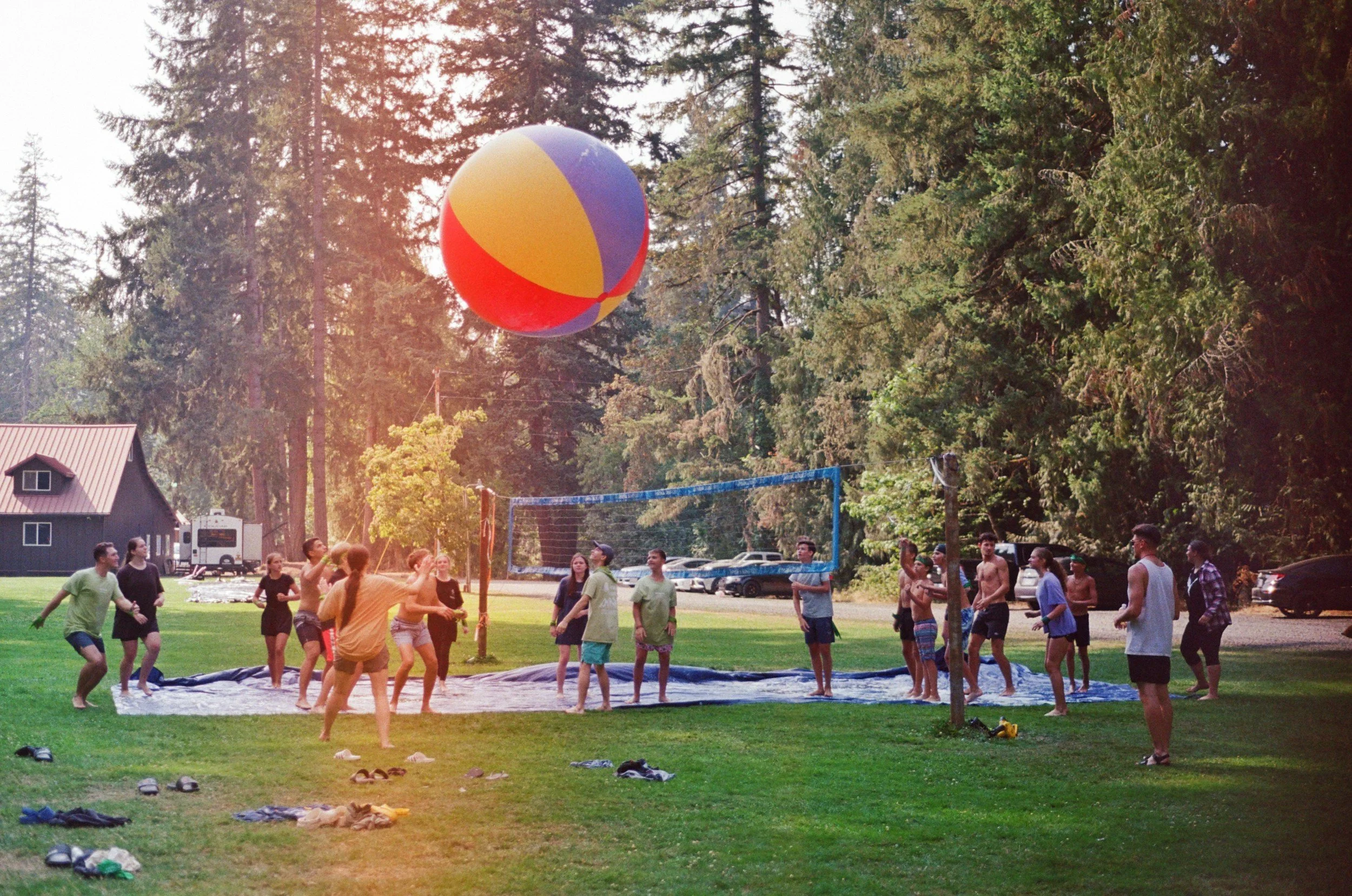 Young people playing volleyball outdoors on a grassy field surrounded by trees, with some shoes and clothing scattered on the ground, and sunlight filtering through the trees.