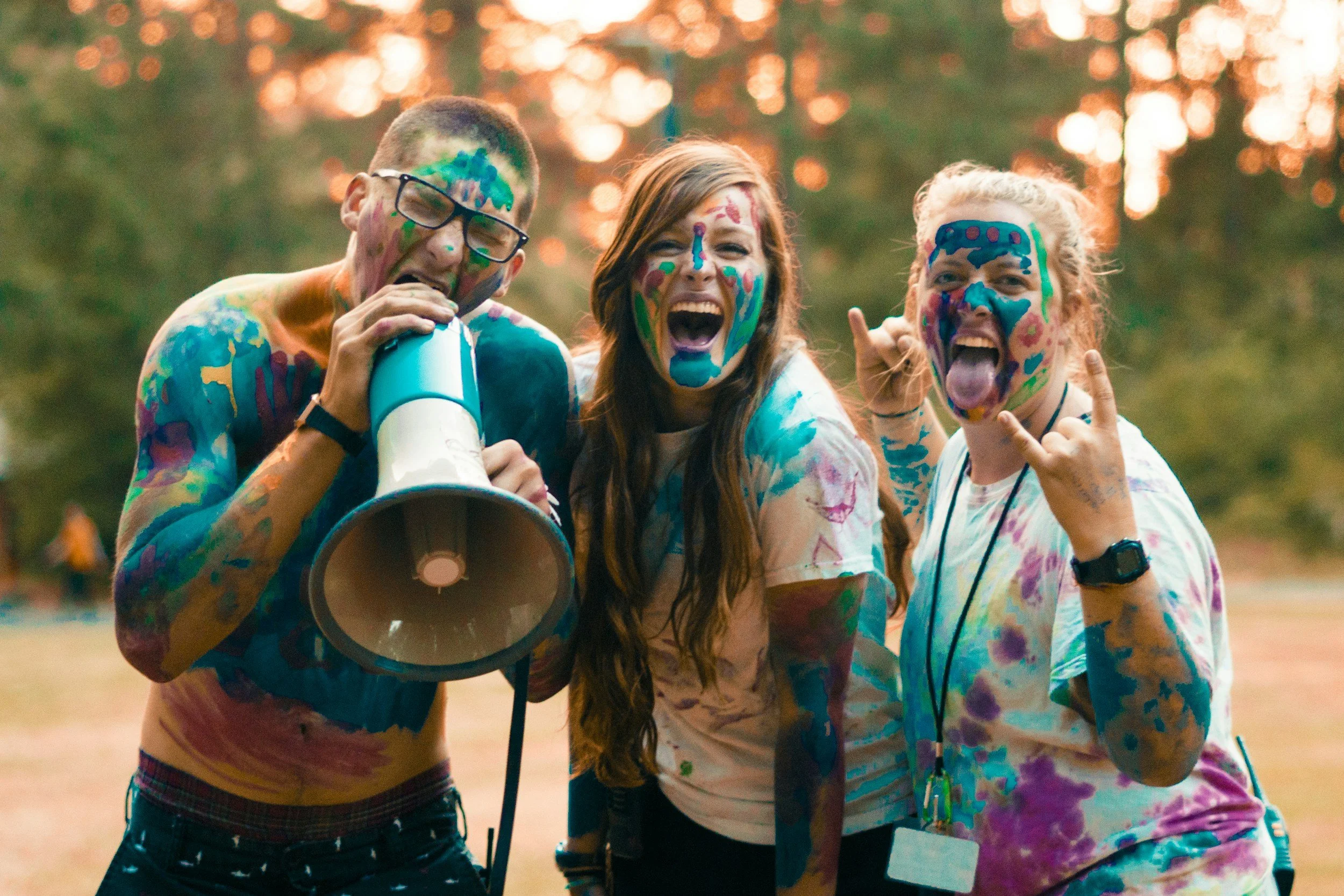 Three young people covered in colorful paint, smiling and having fun outdoors during sunset.