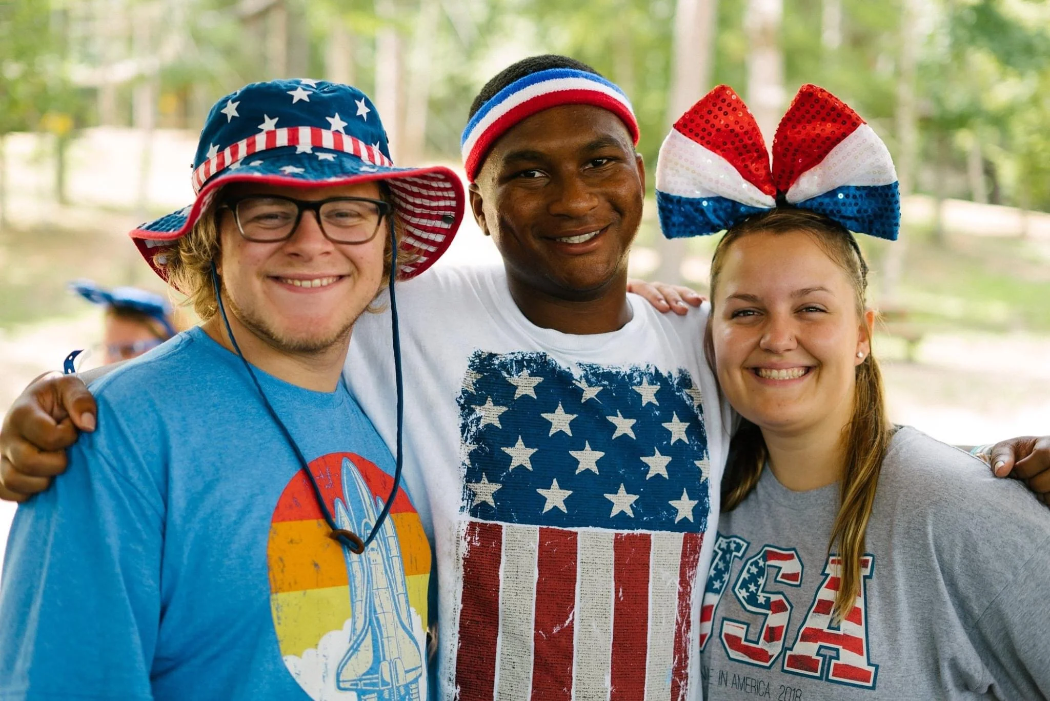 Three young people smiling and standing close together outdoors, wearing patriotic American-themed clothing and accessories, with a lush green forest background.