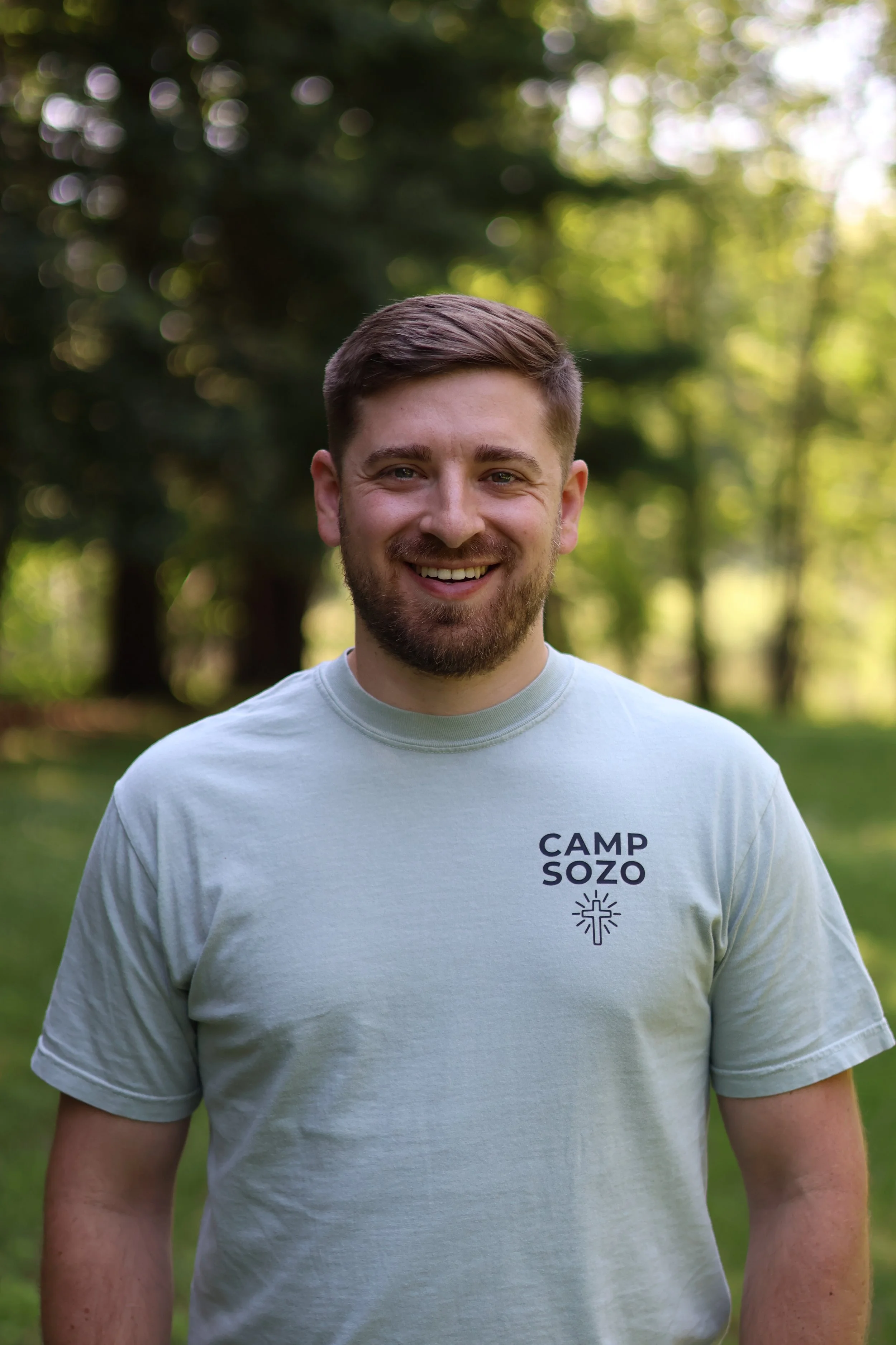 A young man with brown hair and a beard smiling outdoors in a lush green park, wearing a light gray shirt with "CAMP SOZO" printed on it.
