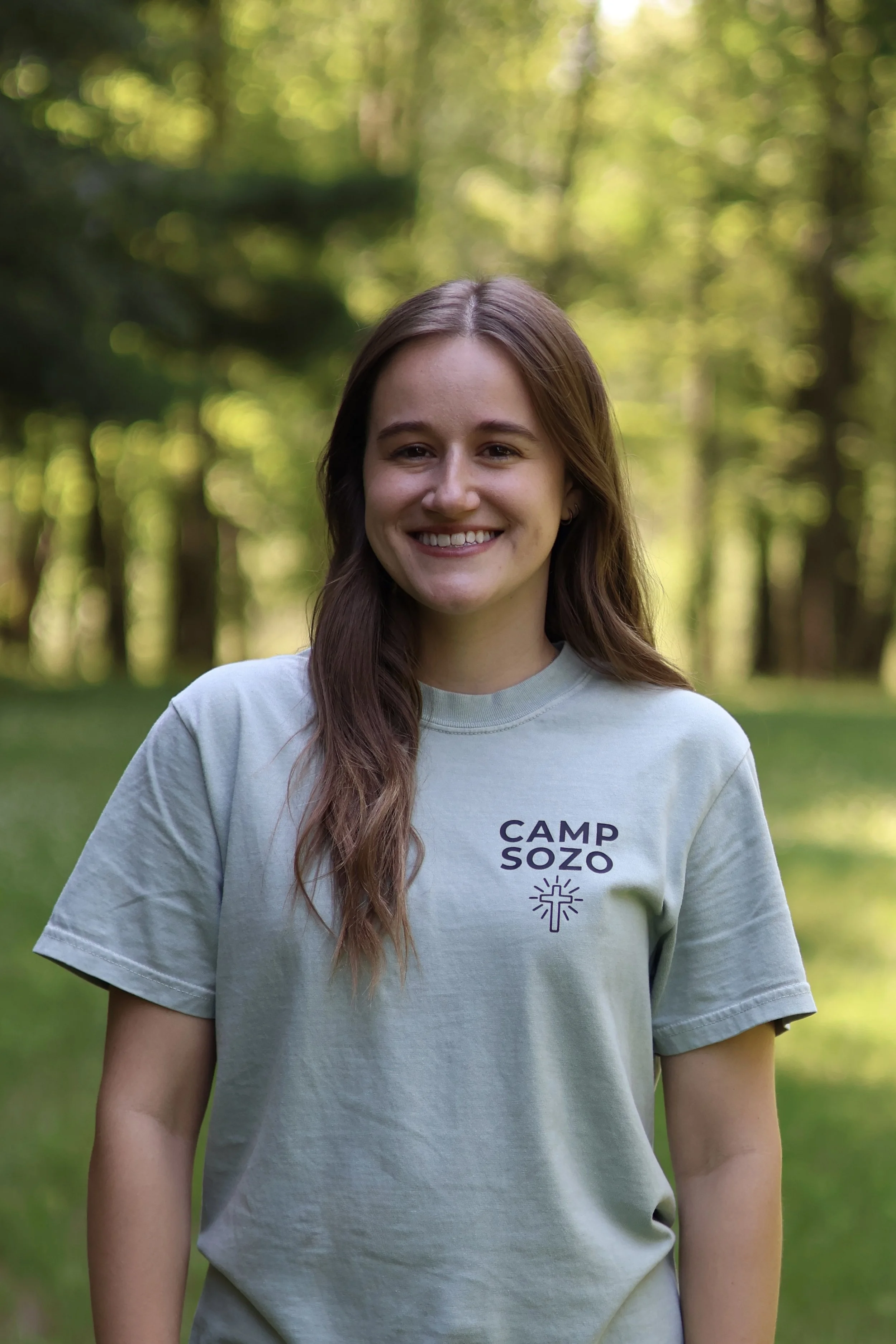 A young woman with long brown hair, smiling, wearing a light gray T-shirt with