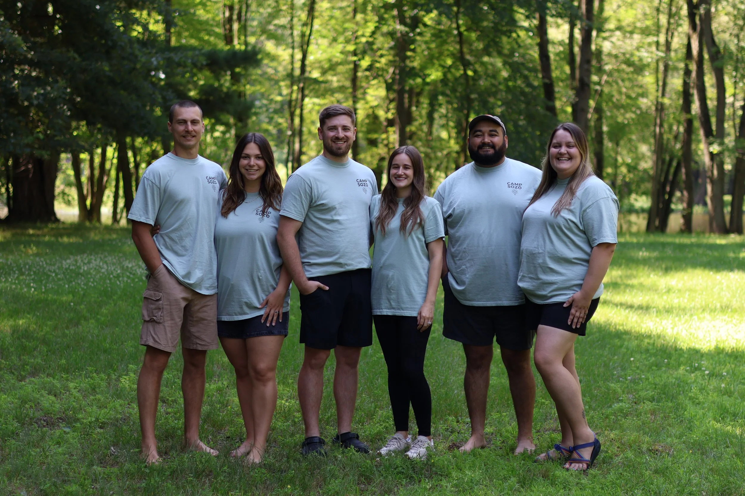 Group of six young adults outdoors in a wooded area, smiling and standing close together, wearing light-colored t-shirts with a camp logo.