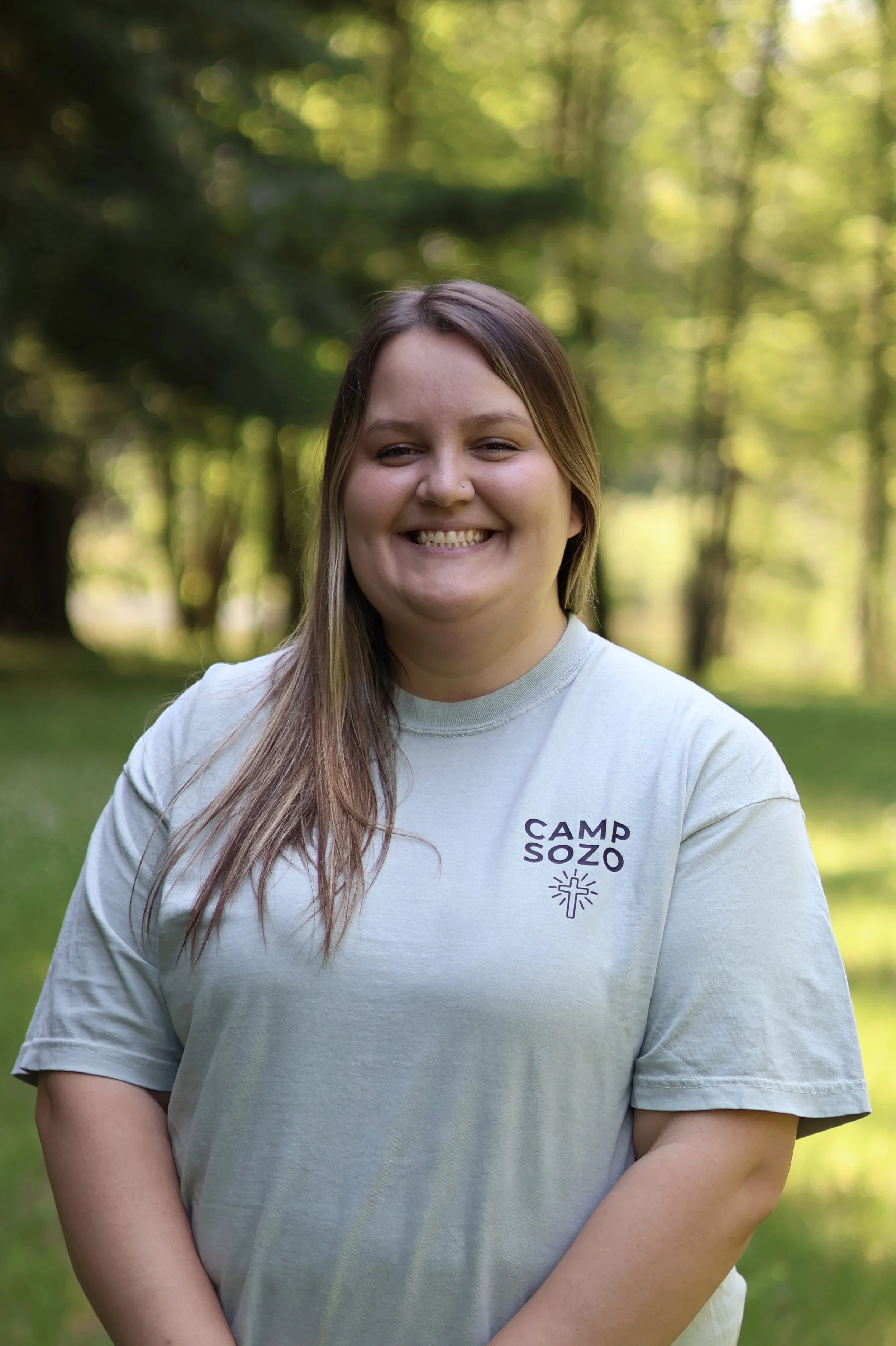 A young woman smiling outdoors in a forest setting, wearing a light gray t-shirt with the words 'Camp Sozo' and a small graphic on it.