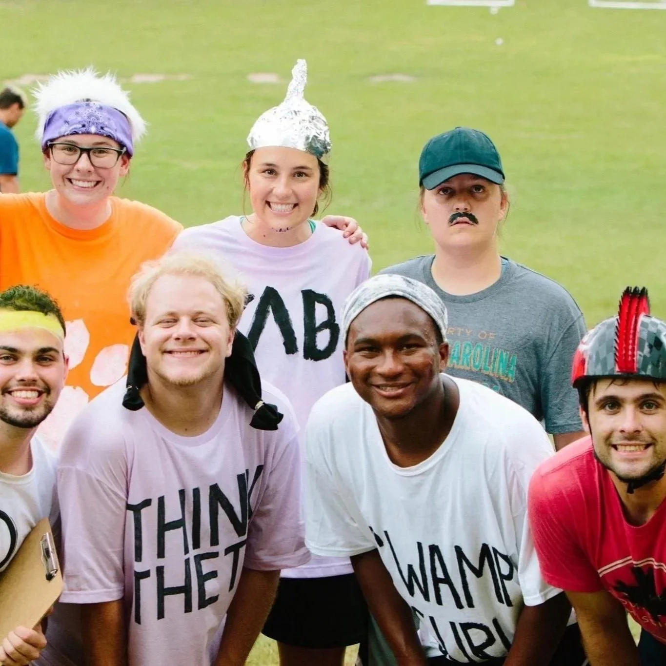 Group of people wearing themed costumes and t-shirts on a grassy field, smiling for the camera during a fun outdoor event.