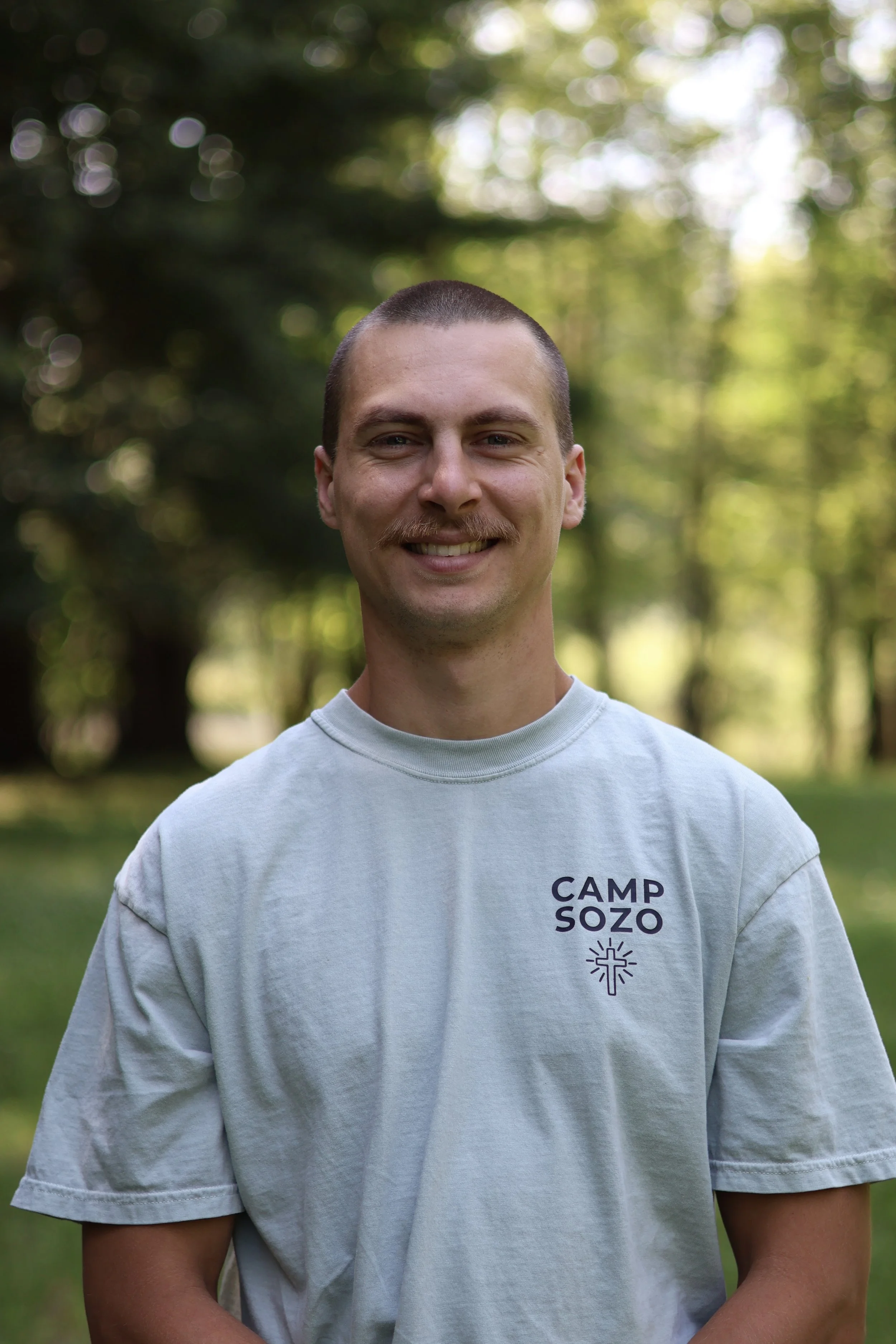 A young man with short dark hair and a mustache smiling outdoors, wearing a light gray T-shirt with purple text that reads 'CAMP SOZO' and a small purple cross and sun design below the text.