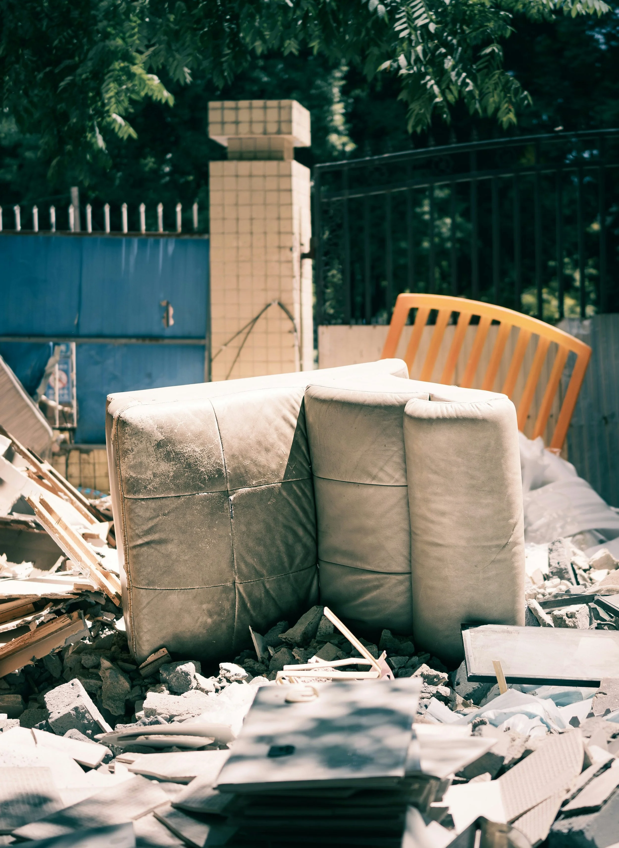 An outdoor scene with a piece of furniture, possibly a cushioned armchair, amidst rubble and debris, with a driveway gate, a brick pillar, and trees in the background.