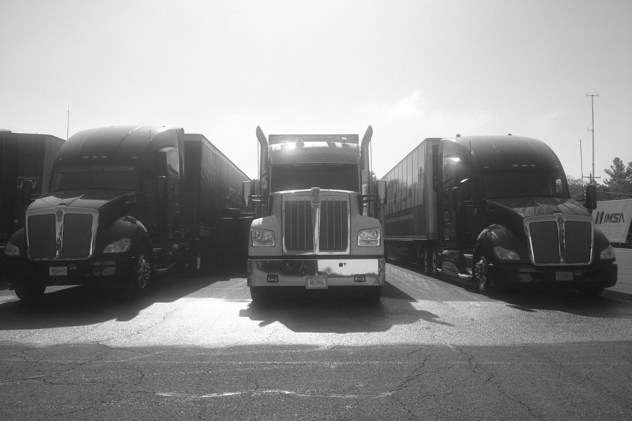 Lineup of four trucks parked in a lot, with two cars partially visible at the edges, and trees and a utility pole in the background, under a cloudy sky.