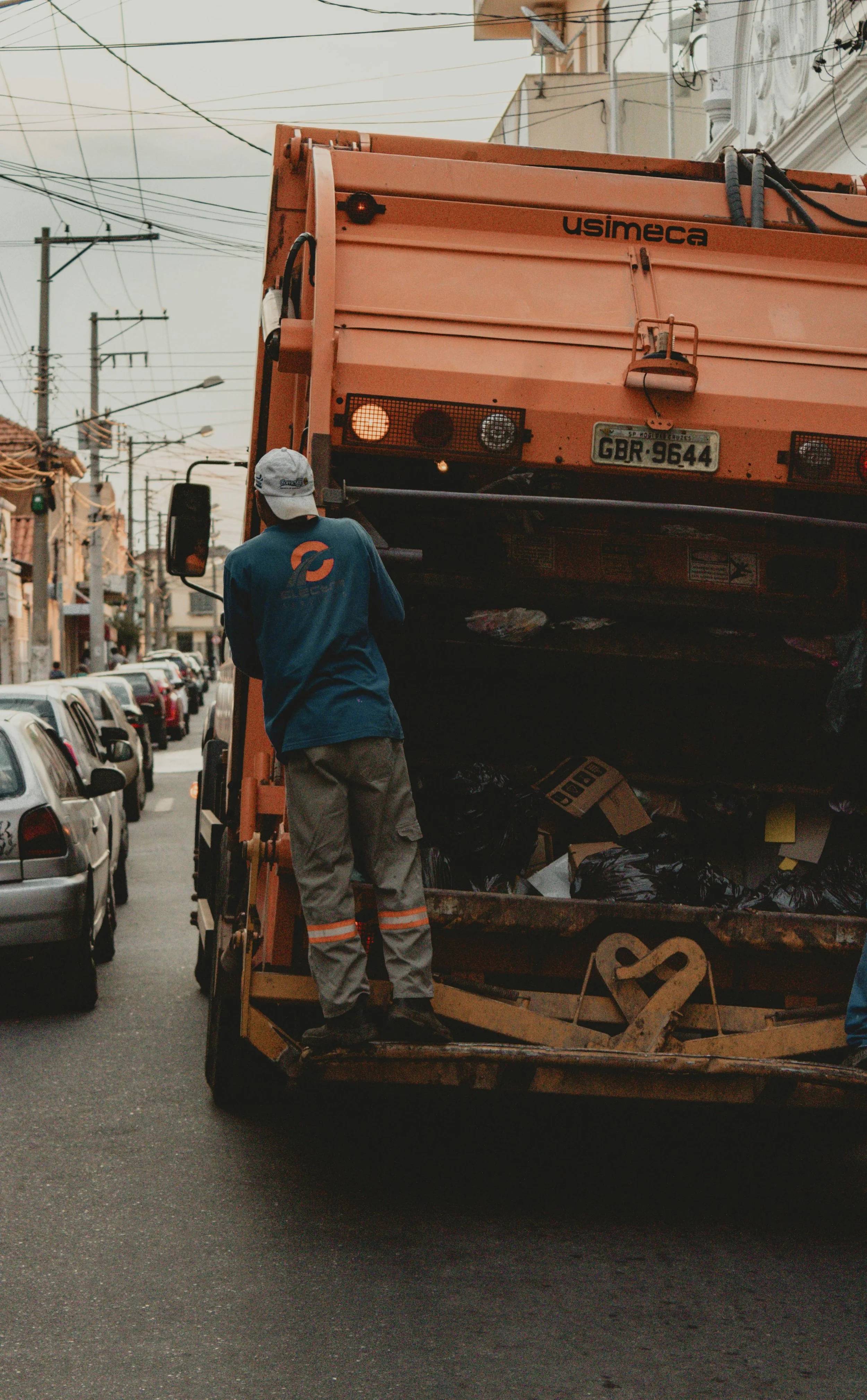 A worker in a uniform and cap standing on the back of a trash collection truck, placing black trash bags into the truck, on a city street lined with parked cars. Overhead power lines are visible, with buildings and cloudy sky in the background.