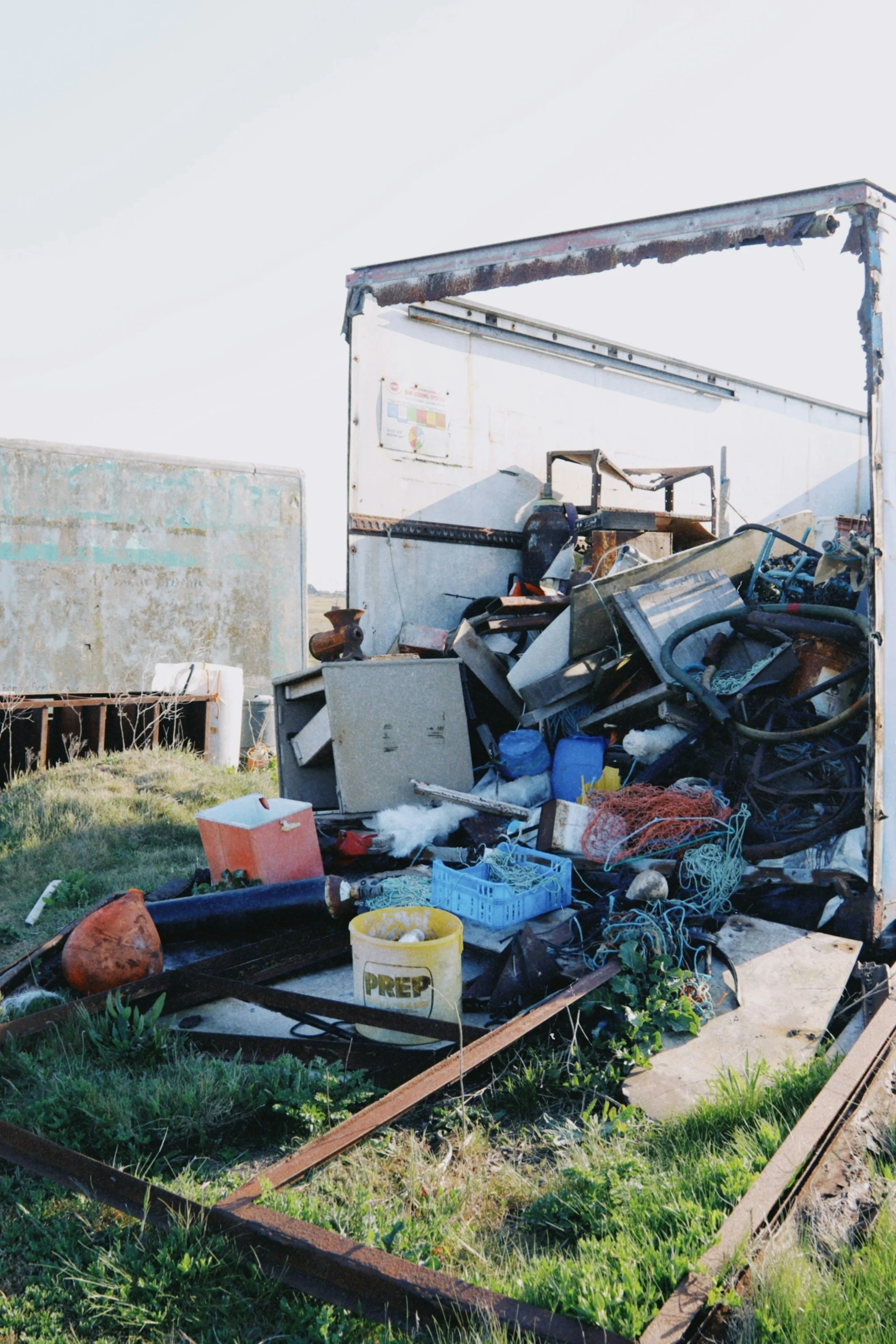 A pile of discarded items and debris outside on a sunny day, including metal, plastic, and wood. There are old appliances, containers, wires, and other miscellaneous trash scattered on the ground with a grass area in the foreground.